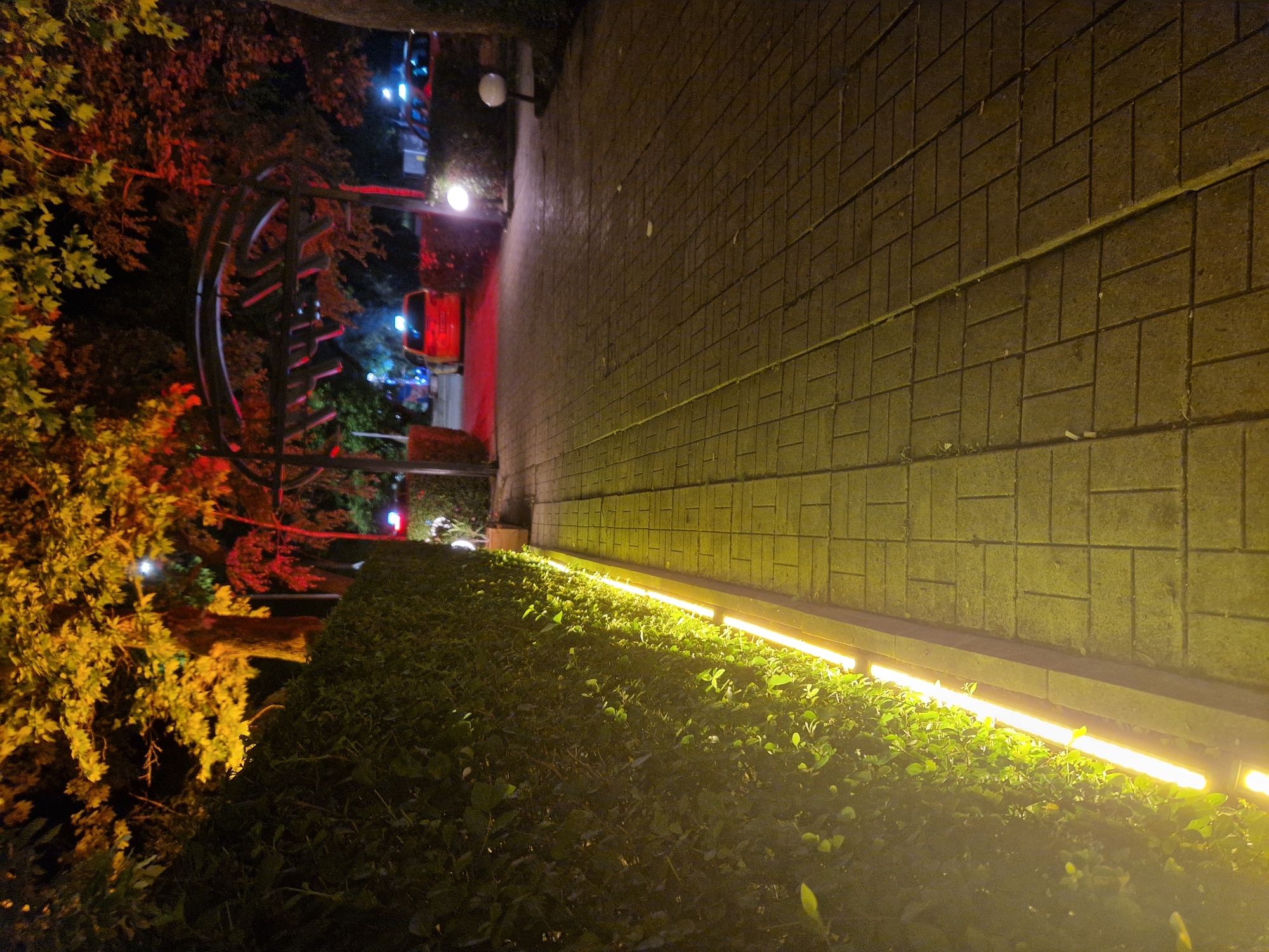 The image displays an outdoor scene at night, illuminated by various artificial light sources. A patterned wall, constructed from rectangular blocks or bricks, extends diagonally from the mid-left towards the upper right of the frame. Below this wall, a series of bright yellow strip lights are mounted, casting a strong, warm glow onto the lower section of the wall and a dense, dark green hedge situated in the bottom-left portion of the image. The hedge runs parallel to the wall and the strip lights.
In the background, to the left of the wall and behind the hedge, are trees and bushes. Some foliage is strikingly lit with a vivid red light, contrasting with the dominant yellow illumination. Amidst these trees, a large, dark, circular or semicircular metal structure, possibly a piece of art or garden architecture, is visible. Further in the distance, bright points of white and blue light can be seen, likely originating from distant streetlights or vehicles. A spherical white lamp on a pole is also present near the wall in the mid-ground.
The time of day is clearly night, indicated by the darkness and reliance on artificial lighting. There are no people or discernible activities taking place, creating a static and tranquil atmosphere. The prominent textures of the block wall and the dense foliage are highlighted by the strong lighting. No text is visible anywhere in the image.