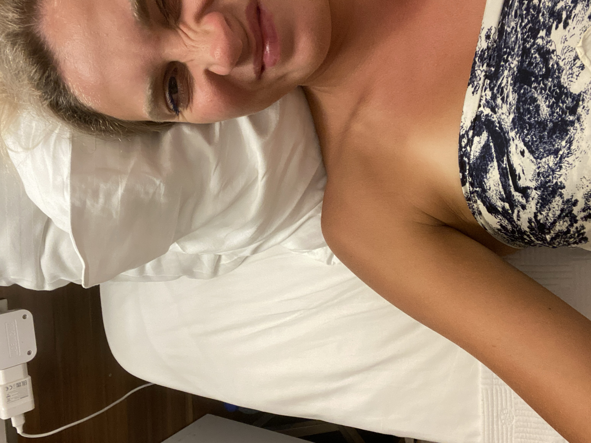 A woman with blonde hair is lying on a white bed. She is wearing a strapless top with a dark blue and white pattern. Her right arm is extended and resting on the white sheet. She is looking towards the camera and making a scrunched-up face. Her right eye has blue eyeliner. In the background, a white power adapter is plugged into an outlet. The scene appears to be indoors.