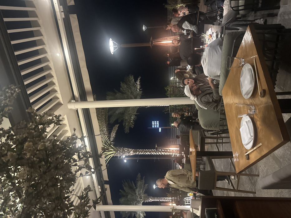 An outdoor restaurant scene at night. Guests are seated at wooden tables with plates and chopsticks. Some individuals wear traditional Middle Eastern attire. Palm trees and streetlights are visible in the background. The area is covered by a partially open roof structure, decorated with lights and what appears to be white flowering plants. The scene suggests an upscale, modern establishment.