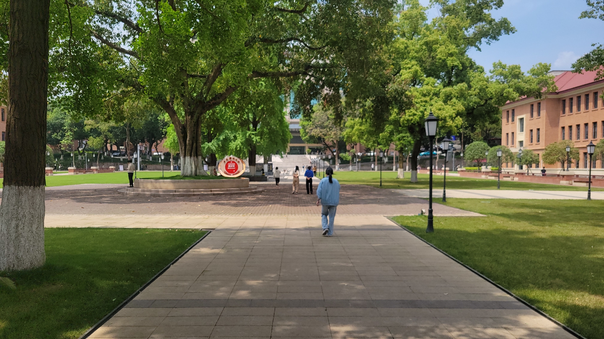 The image shows a wide paved pathway in an outdoor setting. A person in a blue outfit walks away from the viewer along the path. Several other people are visible further down the path. The pathway is flanked by grassy areas and trees, with brick buildings in the background. There are also numerous lampposts lining the sides of the pathway. A large sign is located near the center of the scene. The image is taken during the daytime, with ample sunlight. The location is unknown.