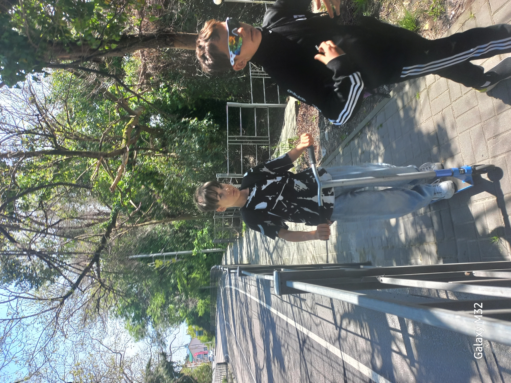 The image shows two people in an outdoor setting. One person stands on the sidewalk beside a silver metal guardrail. This person has dark hair and wears a black and white patterned shirt, jeans, and white sneakers. The person's hands grip the handlebar of a silver and blue pogo stick. This person looks toward the opposite end of the guardrail.
The second person is nearby, near the top edge of the image. This person wears a black Adidas tracksuit and colorful sunglasses. The ground beneath the second person appears to be paved with gray blocks.
The environment includes green trees. A road stretches into the distance beyond the guardrail. The image seems to be taken on a sunny day. The location is Sochi, Russia.