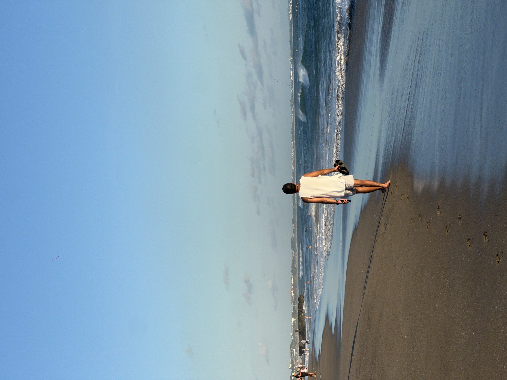 A man in a white shirt and shorts is walking barefoot on a beach. He's holding his sandals in his right hand. The beach has wet sand near the shoreline and drier sand further up. There are footprints in the sand. The ocean waves are visible next to the beach. In the distance, there are buildings and other people on the beach. The sky is blue with some clouds. The location is North Kuta, Indonesia.
