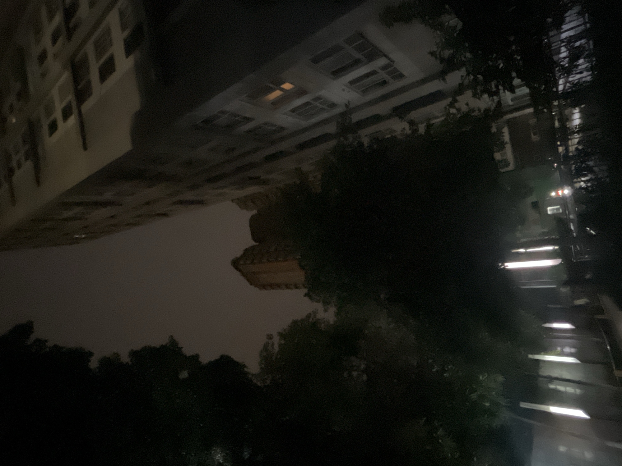 A night-time, low-angle outdoor shot captures a street scene in Jiulongpo District, China. The foreground is dominated by dark, silhouetted trees, with vertical light fixtures casting bright, elongated beams onto what appears to be a pathway or street. In the mid-ground, another building, possibly residential, is visible, illuminated with a warm light from a single window. To the right, a tall, modern building looms, its facade showing multiple windows, some with a faint orange glow, suggesting interior lights. The sky is overcast and dark, contributing to the overall dim illumination of the scene. The perspective is skewed, with the buildings appearing to lean into the frame.