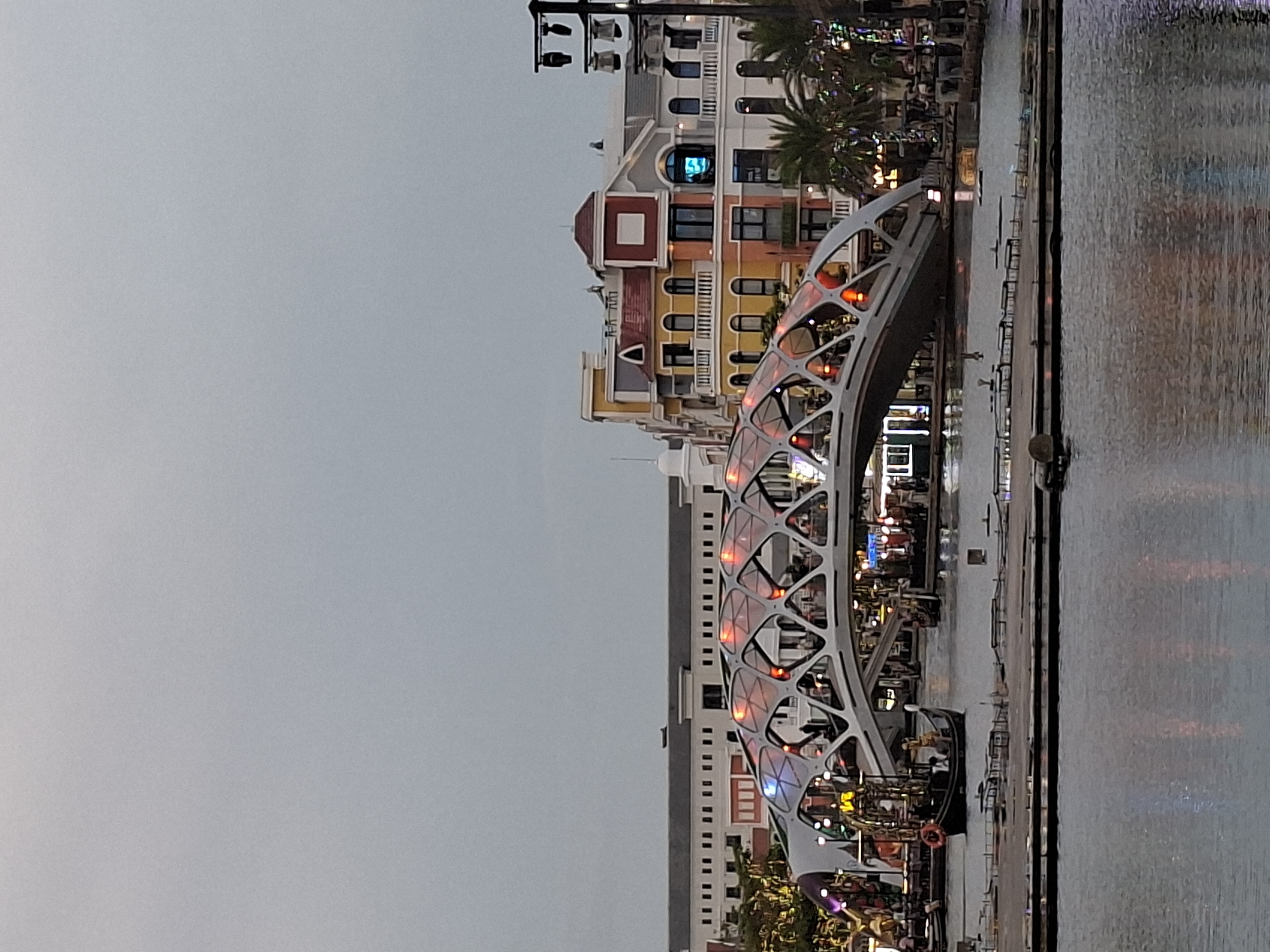 In the city of Phú Quốc, Vietnam, an architectural marvel, a modern bridge with a unique diamond-shaped lattice structure, spans across a body of water. The bridge is illuminated with a mix of red and blue lights, creating a festive atmosphere. Below the bridge, a dark-colored boat, resembling a gondola, navigates the water, carrying a few passengers. On the right side of the image, a row of colorful buildings, with a distinctly European architectural style, stands tall. The buildings are adorned with balconies and large windows, and some are lit up, suggesting commercial activity within. Palm trees are scattered throughout the scene, adding a tropical touch to the urban landscape. The sky is overcast, casting a diffused light over the entire scene. In the foreground, the calm water reflects the lights and structures, creating a mirrored image of the illuminated bridge and buildings. The overall impression is one of a vibrant and dynamic urban setting, blending modern design with classic charm.