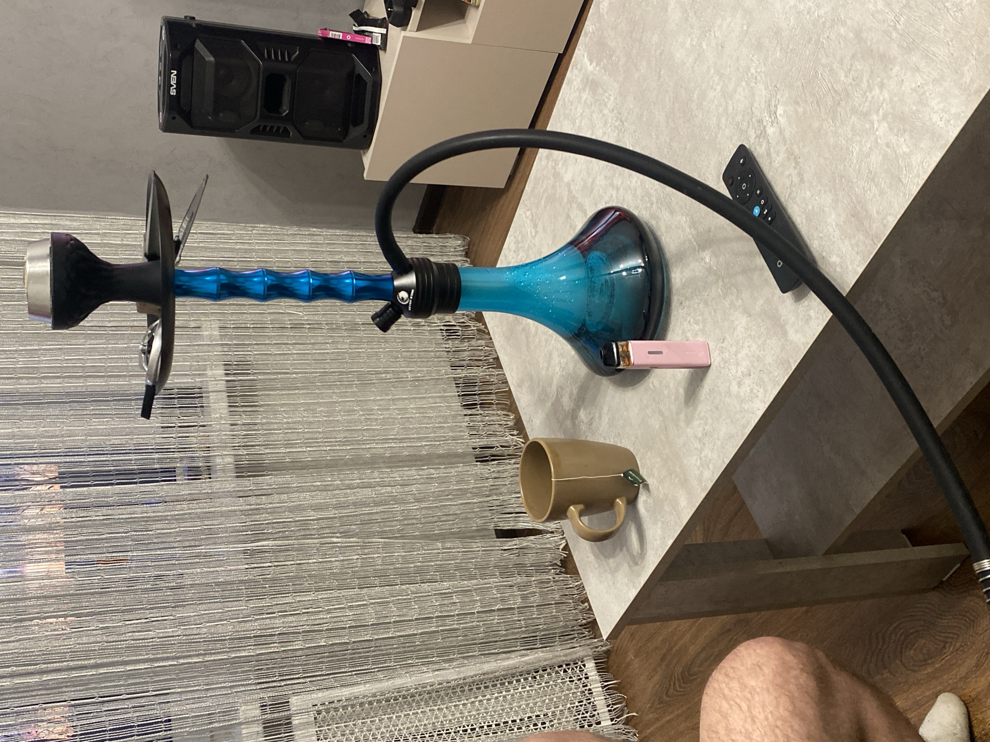 A hookah is centrally located on a table, with a black mouthpiece, blue-tinted central chamber, and a black hose looping around a remote control on the table. A brown mug with a tea bag sits nearby. The table is positioned next to a white cabinet and in front of a curtain made of thin, vertical strings. In the bottom right corner, a person's leg and sock are visible. The city of Izhevsk, Russia, is not visually confirmed.