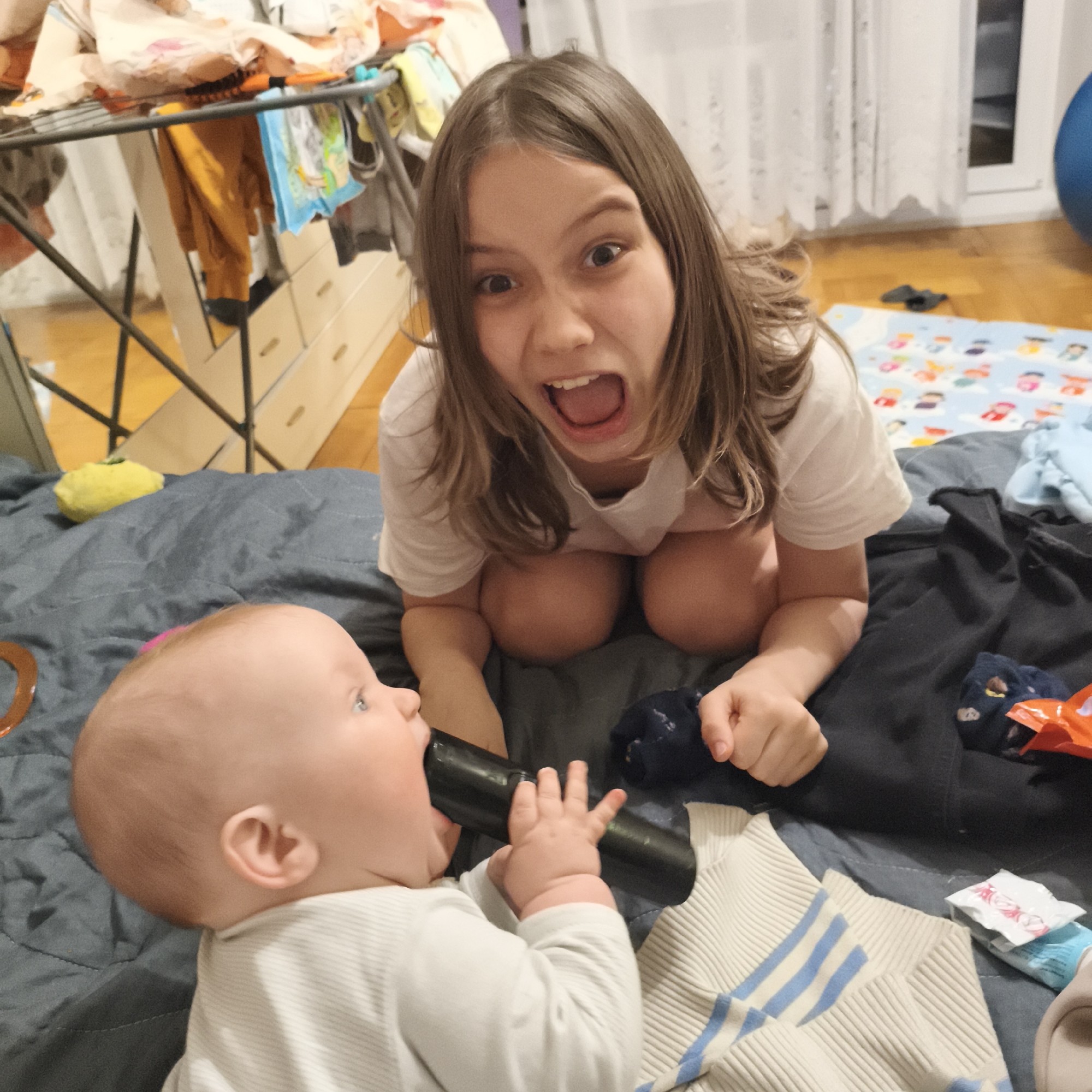 The image shows a young girl kneeling behind an infant who is lying on their back and chewing on a black object, likely a remote control. The girl is making a wide-mouthed expression with her eyes open wide. They are on a grey padded surface with assorted clothing items, including a white sweater with blue stripes and a dark-colored sweater, along with socks and other small items. In the background, there is a clothes drying rack with various garments draped over it, and a blue patterned play mat is visible further back. The location appears to be an indoor domestic setting.