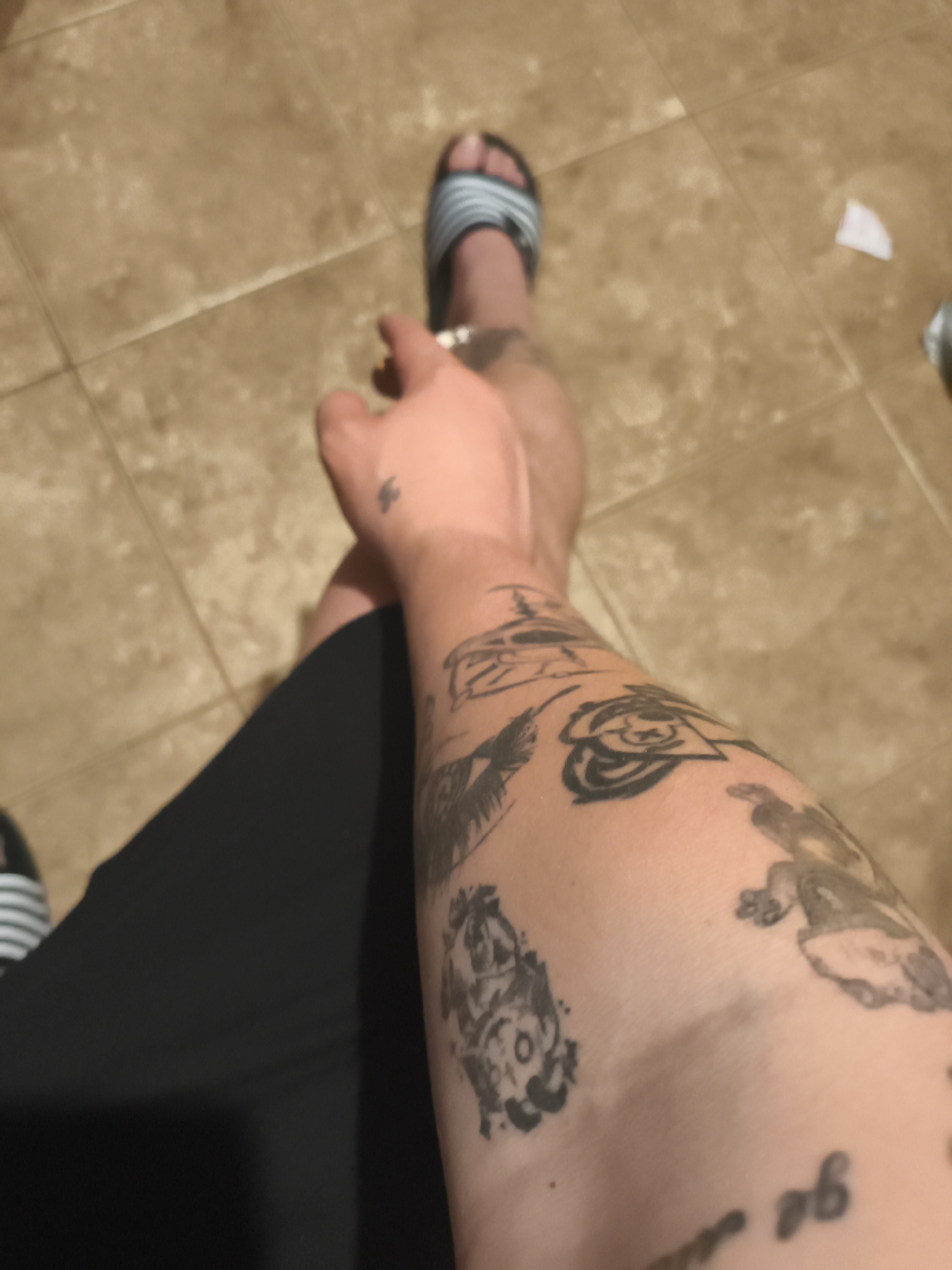 A high-angle, indoor shot depicts a person's tattooed arm and legs. The person is wearing black shorts and striped sandals. The arm has several tattoos of varying designs, including what appears to be a rose, a bird, and cartoon-like figures. The person is holding a lit cigarette in their hand. The background consists of tiled flooring. The image does not contain sufficient information to determine a specific location in Nowy Tomyśl, Poland.