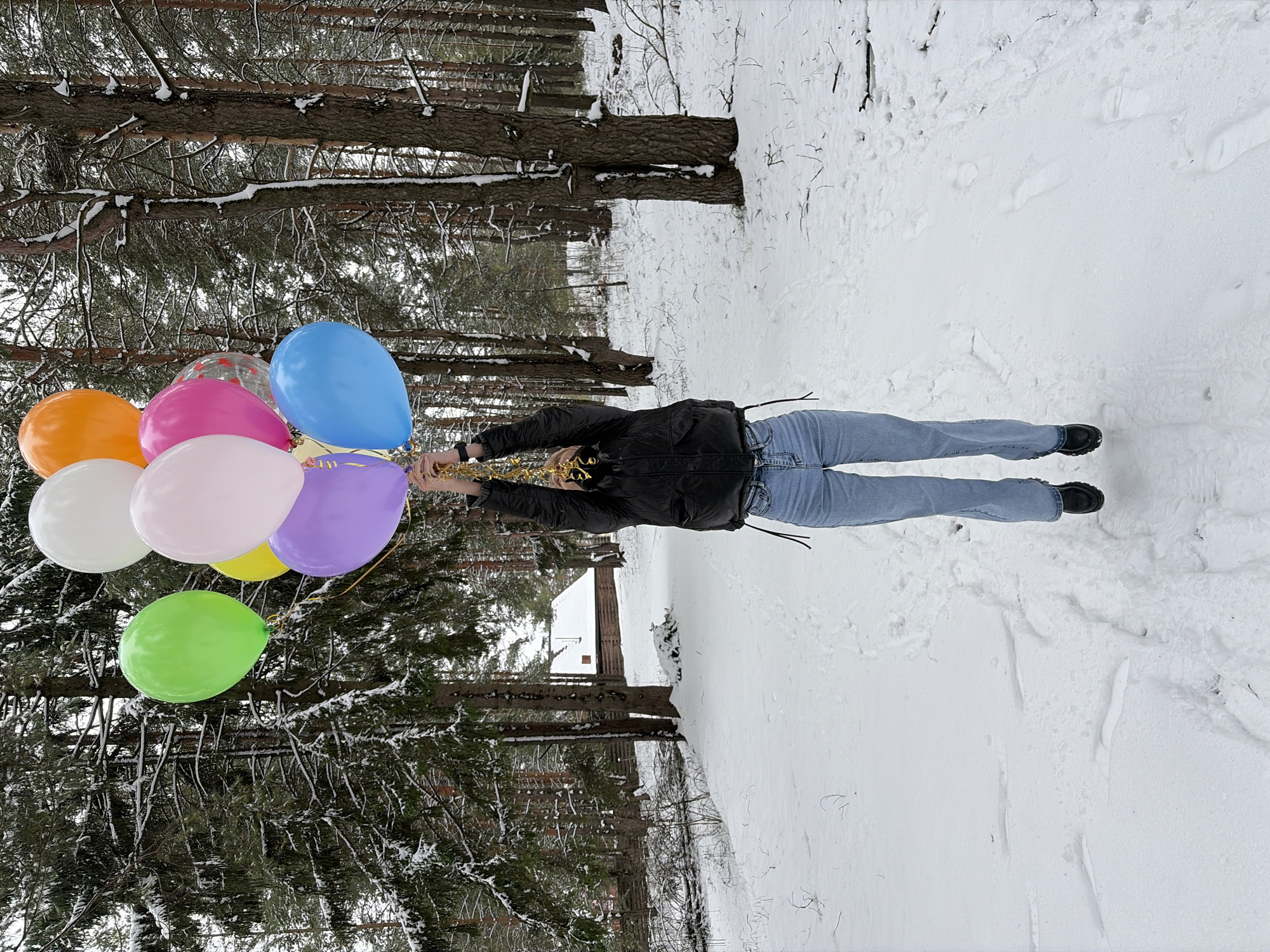 A person is holding a bunch of colorful balloons in a snowy, wooded area. The person is wearing a black jacket and blue jeans, with black shoes. The balloons are various colors including orange, pink, blue, purple, green, yellow, and white. Trees covered in snow surround the person, and the ground is covered in a thick layer of snow. The location is Прикамский, Russia.