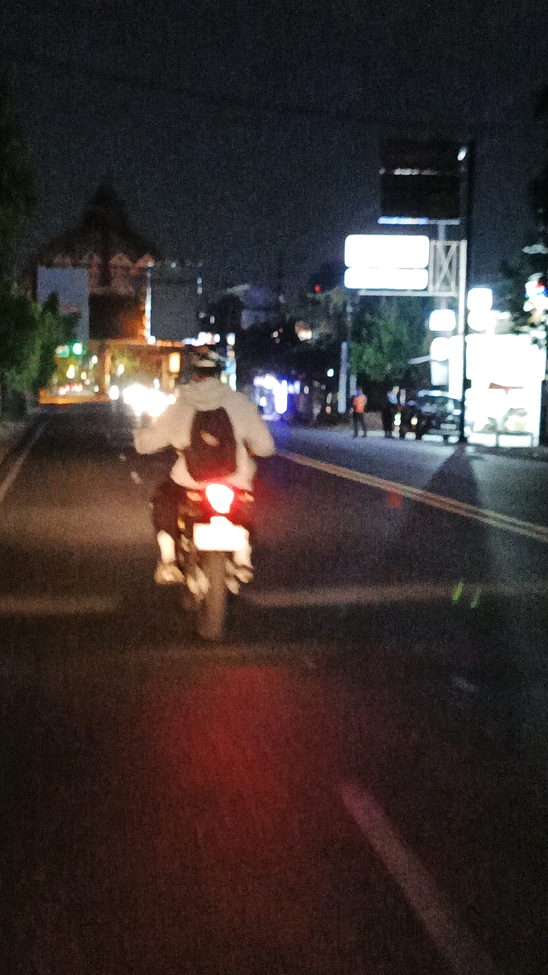 In Blulukan, Indonesia, a person rides a motorcycle at night on a paved road, illuminated by streetlights and vehicle lights. The rider, wearing a white hooded jacket and a backpack, is positioned in the center of the frame, moving away from the viewer. The motorcycle has a lit red taillight and a white license plate. The road has yellow lane dividers. In the background, buildings with illuminated windows and trees are visible, along with street signs and other vehicles. The sky is dark, indicating nighttime.