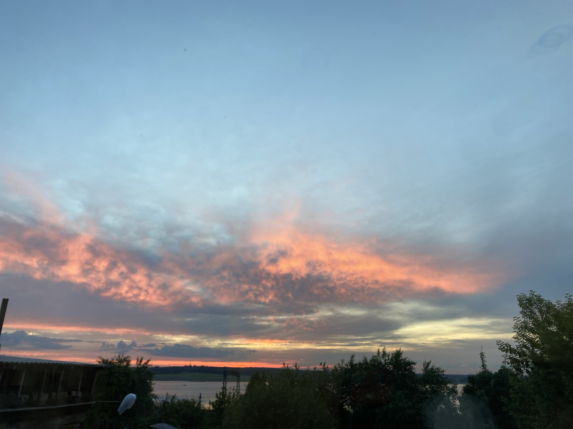 The image presents a serene natural landscape captured at either sunrise or sunset over a body of water in Smolino, Russia. The sky dominates the upper two-thirds of the frame, filled with clouds spectacularly illuminated by the low sun.
The upper portion of the sky is a soft, clear blue, gradually transitioning downwards to lighter grey and yellow tones. A prominent, elongated band of clouds stretches across the mid-sky, glowing with vivid orange, pink, and peach hues. These brightly colored clouds create a striking contrast against the surrounding darker grey and blue cloud formations. Below this, a bright strip of intense orange and yellow light defines the distant horizon, indicating the sun's position just out of view or obscured.
A calm body of water, likely a lake or river, extends across the middle ground, reflecting the subdued light from the twilight sky. The foreground is dominated by the dark silhouettes of dense trees and bushes, suggesting a lush, verdant environment. On the far left, a portion of a dark, rustic structure, possibly a roof or part of a building, is visible. Closer to the water on the left, a partial metal structure, resembling a power line support or pier, can be faintly discerned. A distinct white, oblong object, possibly a light fixture or decorative element, is attached to a pole or branch in the lower-middle left.
Several small, dark specks, likely dust on the camera lens or sensor, are scattered across the upper sky. A faint, larger smudge or streak is also visible in the upper right quadrant, suggesting the image may have been taken through a slightly unclean window. The overall atmosphere is tranquil and picturesque, highlighting the natural beauty of the Russian landscape during twilight. No human activity or visible text is present.