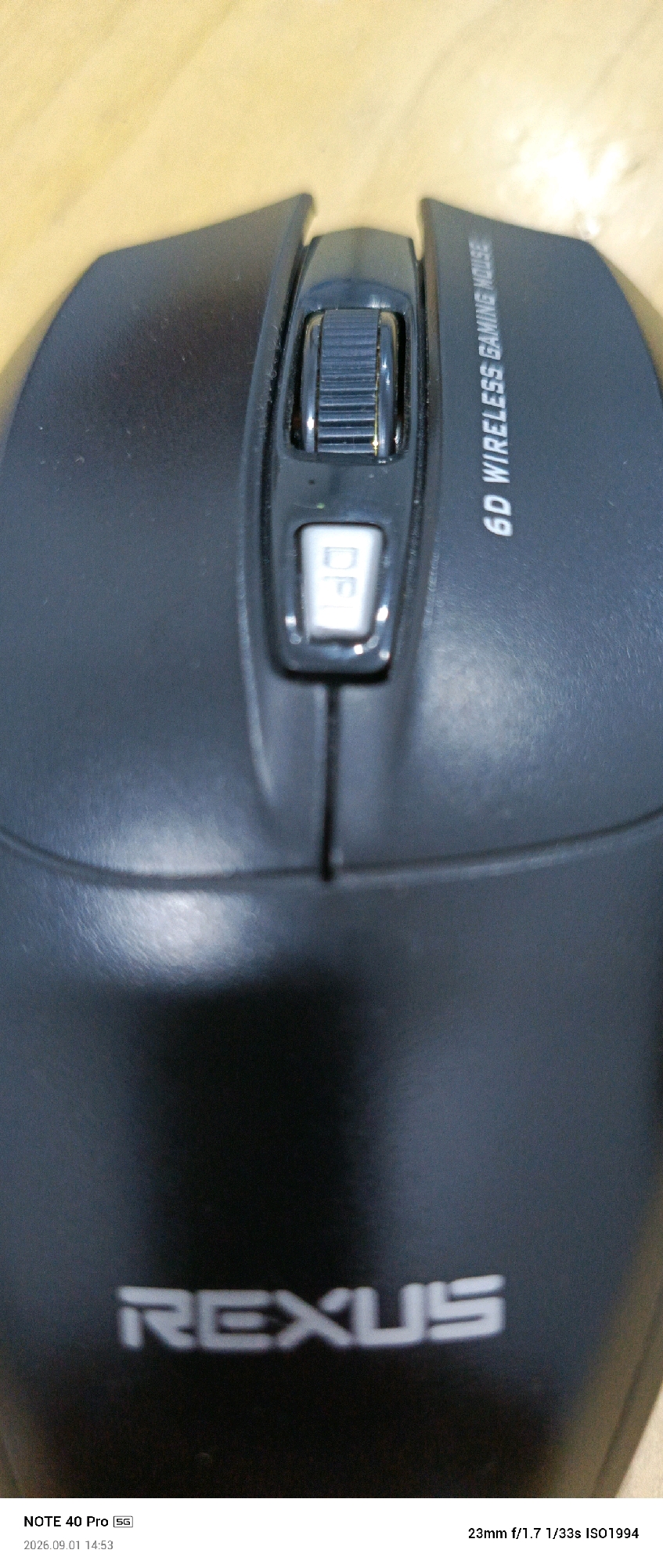 An overhead, medium shot captures a black gaming mouse with a textured scroll wheel and a white DPI button. The mouse is positioned on a light-colored wooden surface. Text on the mouse reads "6D WIRELESS GAMING MOUSE" and "REXUS". A timestamp at the bottom left of the image reads "2026.09.01 14:53". A caption at the bottom left reads "NOTE 40 Pro 5G". A caption at the bottom right reads "23mm f/1.7 1/33s ISO1994". The image appears to be taken in Meunasahaluedrienlhoksukon Barat, Indonesia.