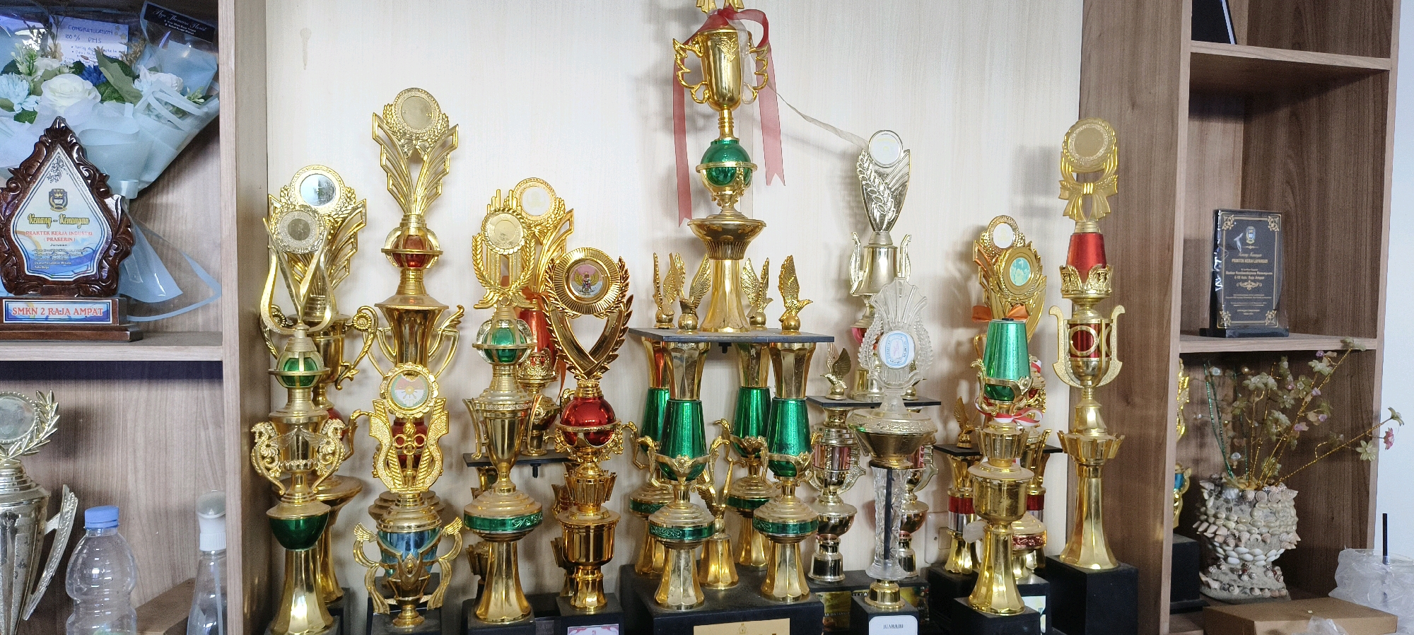 The image shows a collection of trophies and awards displayed on shelves. The trophies are predominantly gold-colored with various embellishments in green and red. Some trophies feature circular medallions with intricate designs. On the left side, a bouquet of white and blue flowers is visible, alongside a framed award with text in Indonesian. The shelves are made of wood, and the background appears to be a light-colored wall. To the right, a wooden cabinet with shelves is also visible, containing a framed award and a decorative vase with artificial flowers. The context of the image being from Waisai, Indonesia, suggests this could be an educational institution or a community center. The scene is static and depicts a display of achievements.