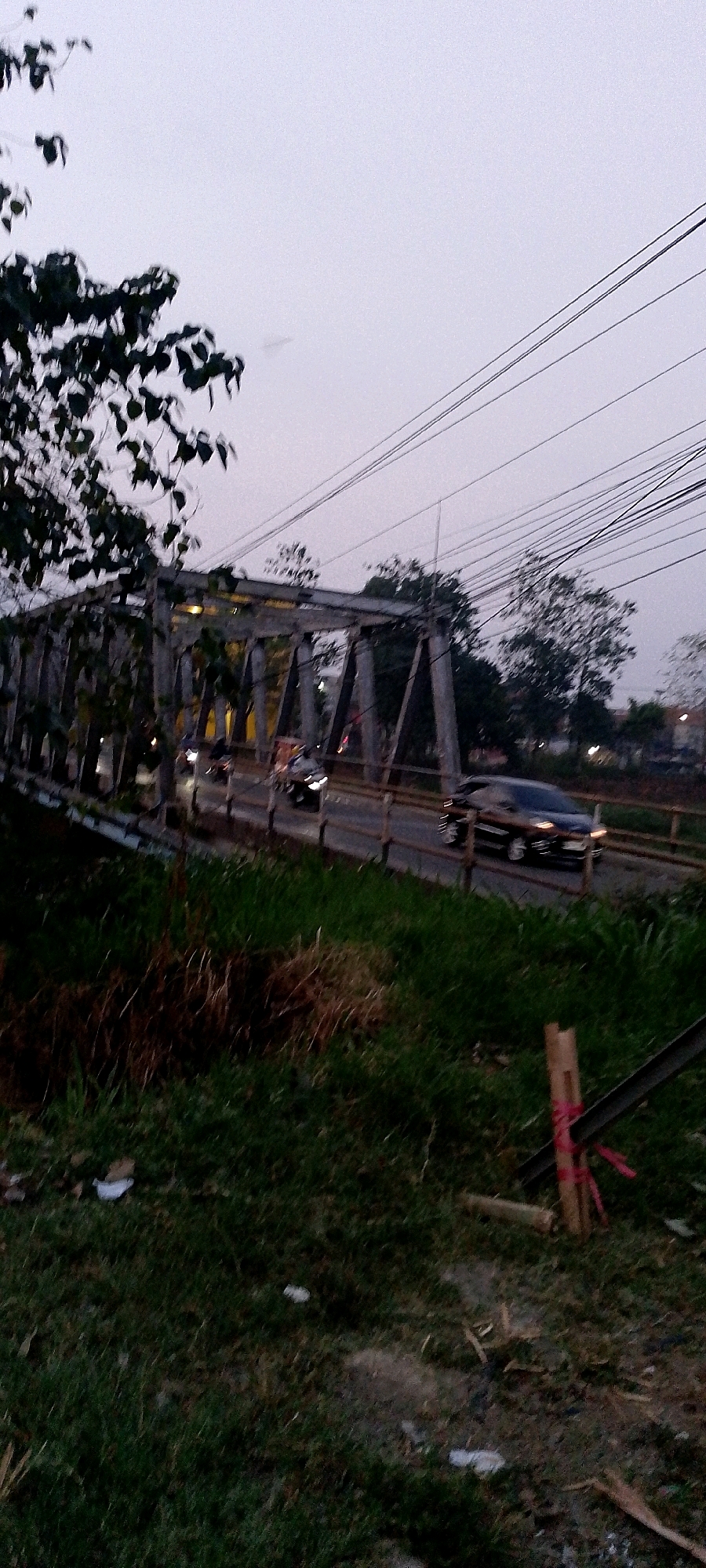 In Margahayu, Indonesia, an overpass bridge is visible as traffic, including cars and motorcycles, moves across it during twilight. The sky is overcast and grey, with power lines crisscrossing the upper portion of the frame. Trees and foliage flank the bridge, with a grassy embankment in the foreground. A metal structure, possibly part of the bridge's construction or maintenance, is present in the lower right corner, with red tape wrapped around it. The lighting suggests it is either early morning or late evening.