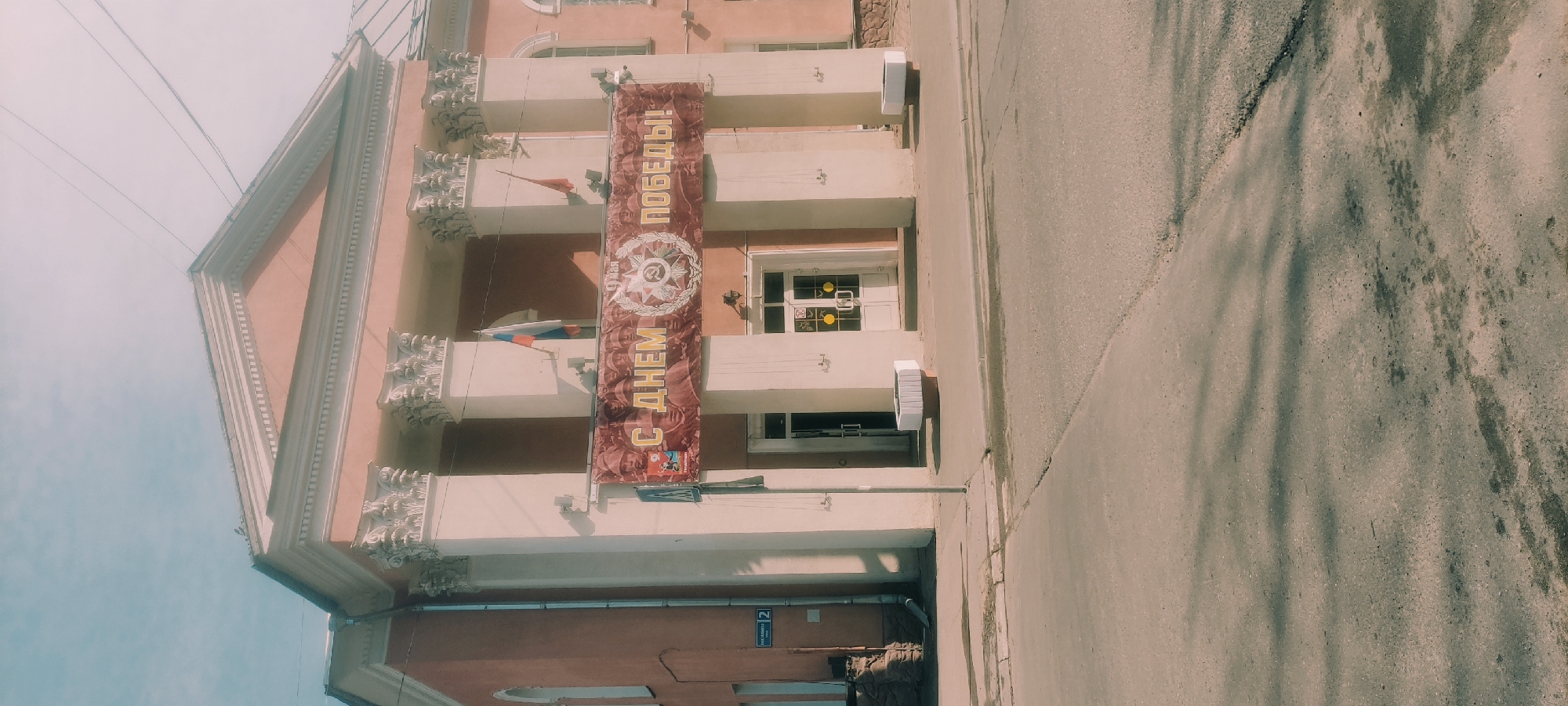 The image shows a three-story building in Fryazino, Russia. A banner hangs vertically on the front of the building displaying the phrase "С ДНЕМ ПОБЕДЫ!", which translates to "Happy Victory Day!". The building features a light pink façade with white architectural details. Russian flags are attached to the balconies. An address plaque reading "2" is visible at the bottom of the building. A sidewalk and street are in the foreground. Overhead electrical wires are visible in the background.
