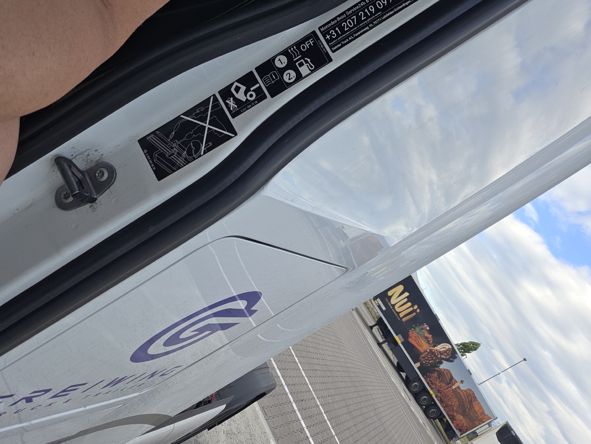 The image shows the interior of a white van, possibly a Mercedes-Benz, with the door open. A person's arm is visible at the top left, with light skin and some arm hair. The van's interior includes a black rubber seal along the door frame and a metal latch mechanism. Several stickers are affixed to the white surface inside the door. One sticker features a diagram of a vehicle with a person, and another shows icons for heat, an open book, and a fuel pump, with the text "OFF" and "1." and "2."
A prominent sticker displays "Mercedes-Benz Service24h" and a phone number "+31 207 219 09". Below this, there is smaller text indicating "Regulieren befonen kunnen in rekening gebracht worden" and "Daimler Truck AG, Fasanenweg 10, 70771 Leinfelden-Echterdingen". Another sticker shows a part number "A 001 584 72 38".
On the exterior of the van, a large purple "G" logo is visible, along with partially visible text that appears to be "GREI/WING TRUCKS".
To the right of the van, a truck trailer is parked, branded with the "Nuii" logo and an advertisement for "ICE CREAM ADVENTURE" featuring Australian landscape imagery. The truck is black with large graphics of ice cream and a desert scene.
The scene appears to be outdoors, with a cloudy sky visible in the reflection on the van's surface and in the background. The ground is paved with a patterned surface, suggesting a parking lot or service area. The overall impression is that this is a daytime shot, likely taken in Flörsheim, Germany, where the Mercedes-Benz truck division is located. The mood is neutral, focusing on details of the vehicle and its branding.