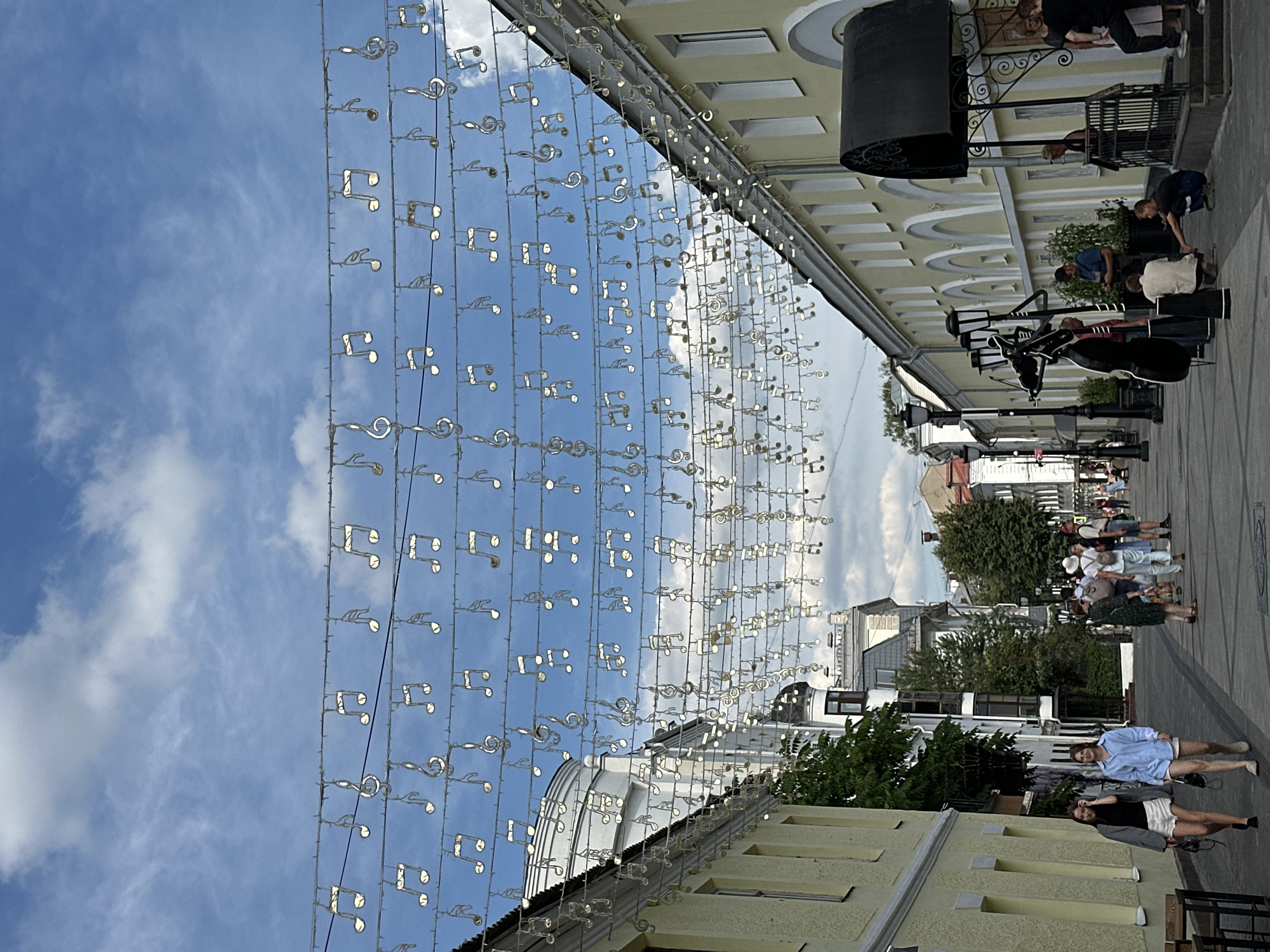 This is a street-level, vertical-format, eye-level shot looking down a pedestrianized street in Putyatino (Semivragovskaya S/A), Russia. The sky is blue with scattered white clouds. Strands of white, illuminated musical notes are strung across the street, decorating the scene. The street is lined with two-story buildings, predominantly yellow with arched window details on the right side. On the left, the building is also yellow with visible windows.
People are walking along the street. In the foreground on the left, two young women walk towards the viewer, one in a gray jacket and white shorts, the other in a blue shirt and white shorts. Further down the street, a group of people are walking in various directions. On the right side of the street, there is a black awning over an entrance, with a railing and stairs. A sculpture of a cello and a figure appears to be on the sidewalk near a lamppost. Several individuals are gathered near this sculpture and the building entrance. The street pavement is visible, and lampposts are present along the sidewalk. Trees are interspersed among the buildings.