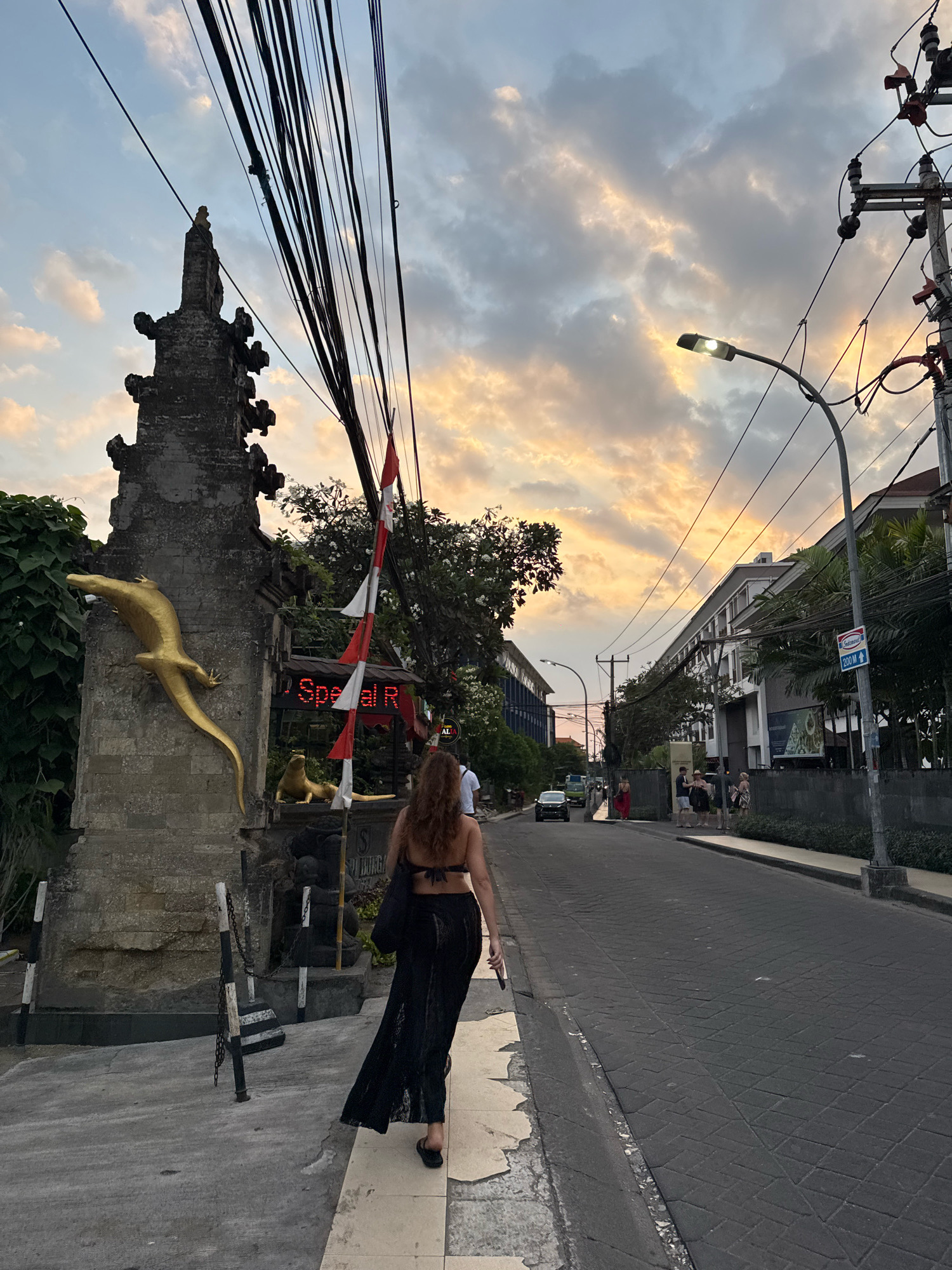 A woman wearing a black bikini top and a long black lace skirt walks away from the camera on a street in Kuta, Indonesia. She is holding a cell phone in her right hand. To her left, a stone gateway is adorned with a golden lizard sculpture and a smaller golden lizard sculpture at its base. Red and white flags fly from the gateway. Behind the gateway, a neon sign with red lettering reads "Special R". A street lamp illuminates the sky, which is filled with dramatic clouds illuminated by the setting sun, casting hues of orange and yellow. The street is paved with dark cobblestones and lined with buildings and trees. Several other pedestrians are visible further down the street, as well as cars and a scooter. Utility poles with numerous black wires crisscross overhead.