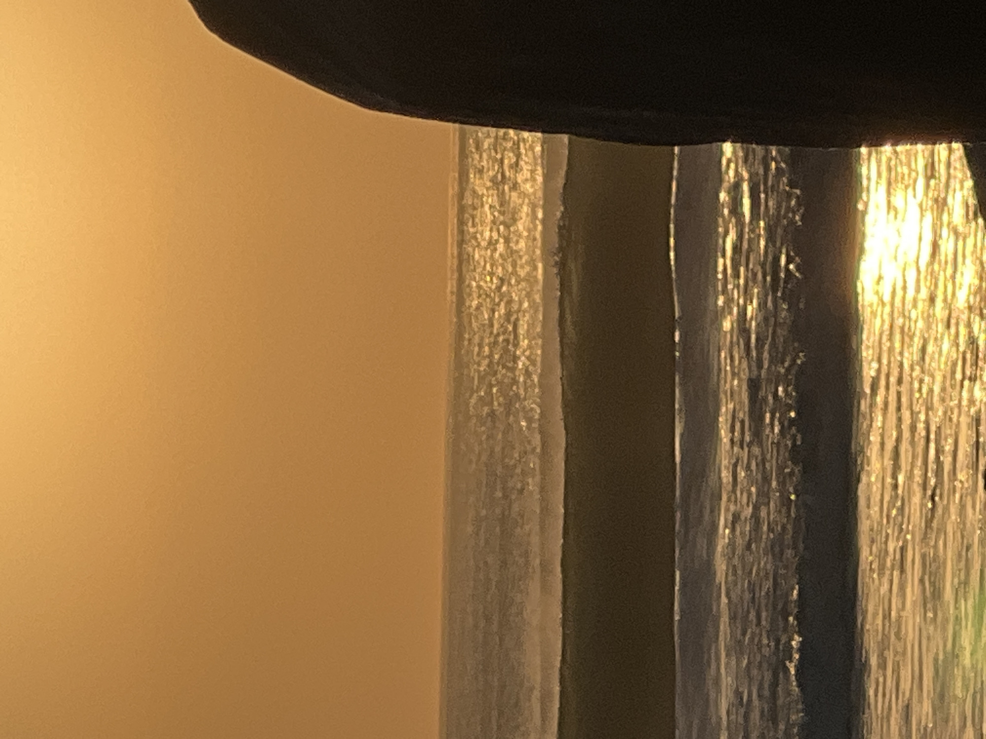 The image shows a silhouette of a person from the shoulders up, seen from the side, standing in the foreground on the right side of the frame. The person is facing left, towards a body of water. The background is dominated by a large expanse of a pale orange sky, suggesting either sunrise or sunset. Below the sky, there are several layers of ocean waves, with the water reflecting the golden light of the sky, creating a shimmering effect. The waves appear to be calm, with no rough seas visible. The environment appears to be a beach or coastal area. The lighting suggests a warm, soft natural light. The overall impression is a serene coastal scene at dusk or dawn. While the image depicts a coastal scene, it is not possible to definitively identify the specific location as Kuta, Indonesia without further visual cues or contextual information.