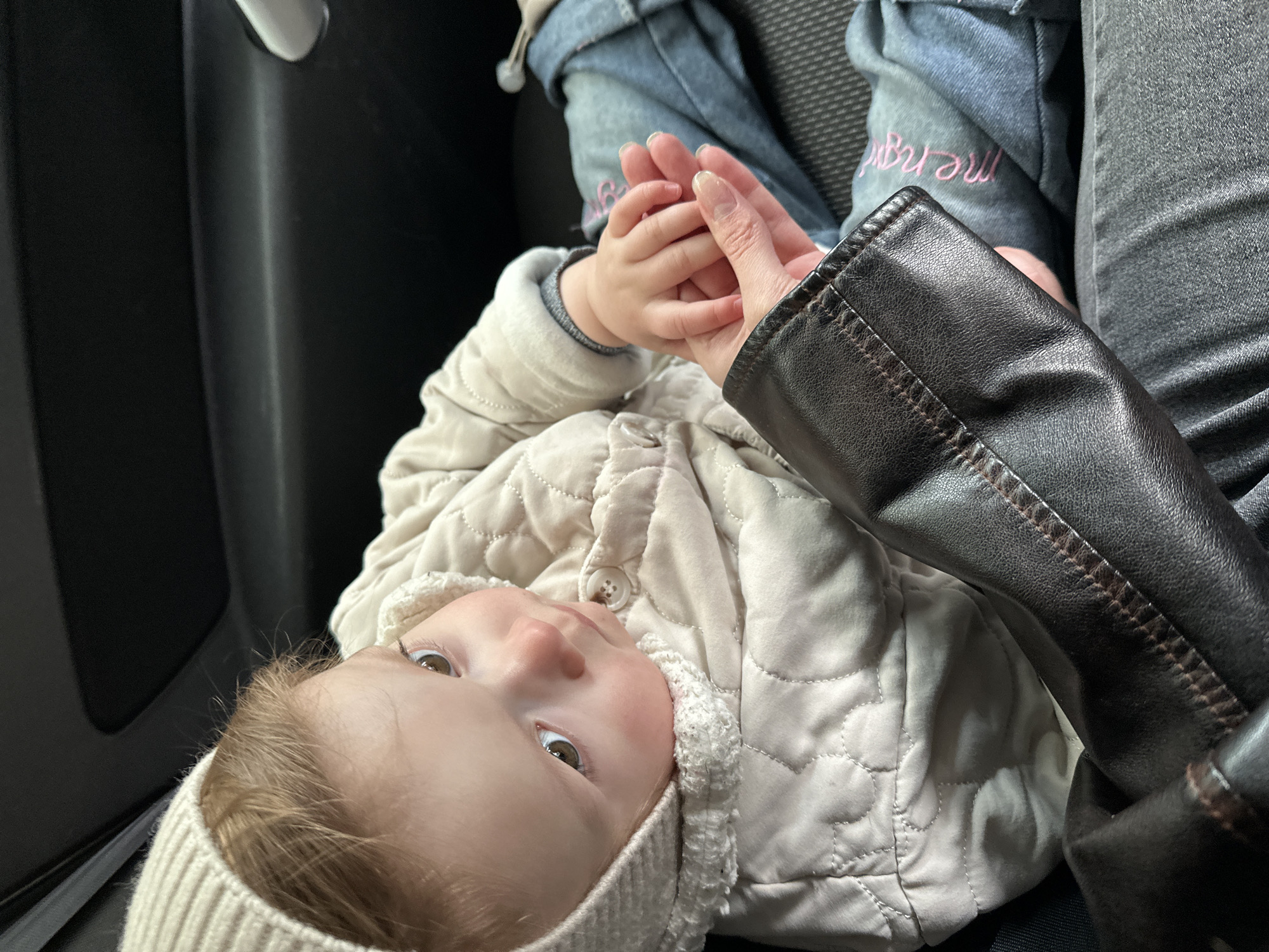 A baby wearing a beige quilted jacket and matching knit hat lies in someone's lap, holding their hand. The person is wearing dark jeans, a denim jacket with pink embroidery, and a black leather jacket. The scene appears to be inside a car. There is no information available in the image regarding the city of Chaikovsky, Russia.