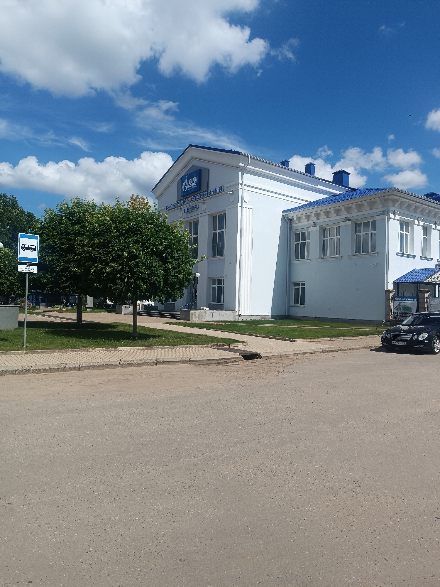 The image depicts a street scene in Chaikovsky, Russia, featuring a large, modern, white building with a blue roof. The building appears to be a public facility, possibly a cultural or sports center, as indicated by the visible signage which includes "ГАЗПРОМ" (Gazprom) and "КУЛЬТУРНО-СПОРТИВНЫЙ ЦЕНТР" (Cultural-Sports Center). The architecture is functional and contemporary, with numerous windows and decorative elements.
In front of the building, there is a well-maintained lawn and two mature trees with rounded canopies, providing a touch of greenery against the urban backdrop. A blue and white bus stop sign is prominently placed on the left side of the image, suggesting this is a public transportation hub.
The street in the foreground is paved with asphalt, showing signs of wear and tear. A black sedan is parked on the right side of the street, partially in front of the building's entrance area. The sky above is a clear, bright blue with scattered white clouds, indicating fair weather. The overall impression is that of a tidy, organized public space within a Russian city.