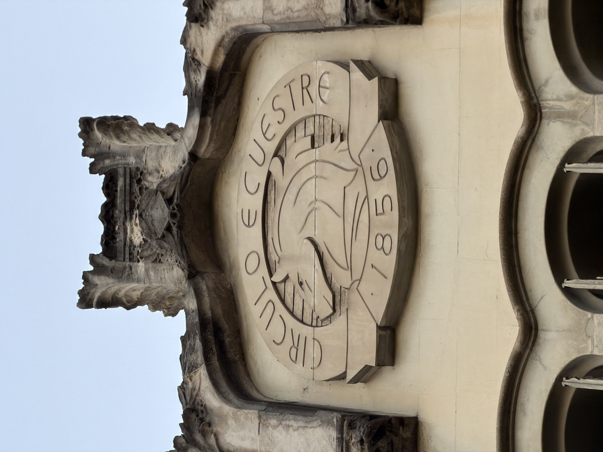 The image shows a relief sculpture on the facade of a building in Barcelona, Spain. The sculpture features a circular emblem with the inscription "CIRCULO ECUESTRE" and the year "1856". Inside the circle, there is a depiction of a horse's head. The sculpture is made of stone and is located above a series of arched windows. The building appears to be old and ornate, with decorative stonework and a weathered appearance. The sky in the background is clear and pale blue. The overall impression is one of historical significance and architectural detail.