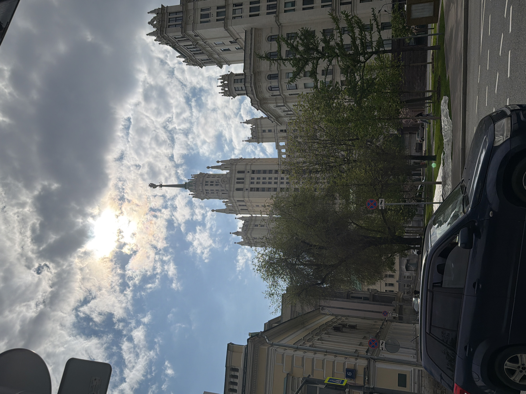 The image shows a view of the Kotelnicheskaya Embankment Building in Moscow, Russia. The building is visible in the upper half of the image, partially obscured by trees. The sky is cloudy with the sun shining through. A dark-colored car is parked on the street in the foreground on the right side of the image. There are other buildings visible in the middle ground on the bottom half of the image and street signs near them.