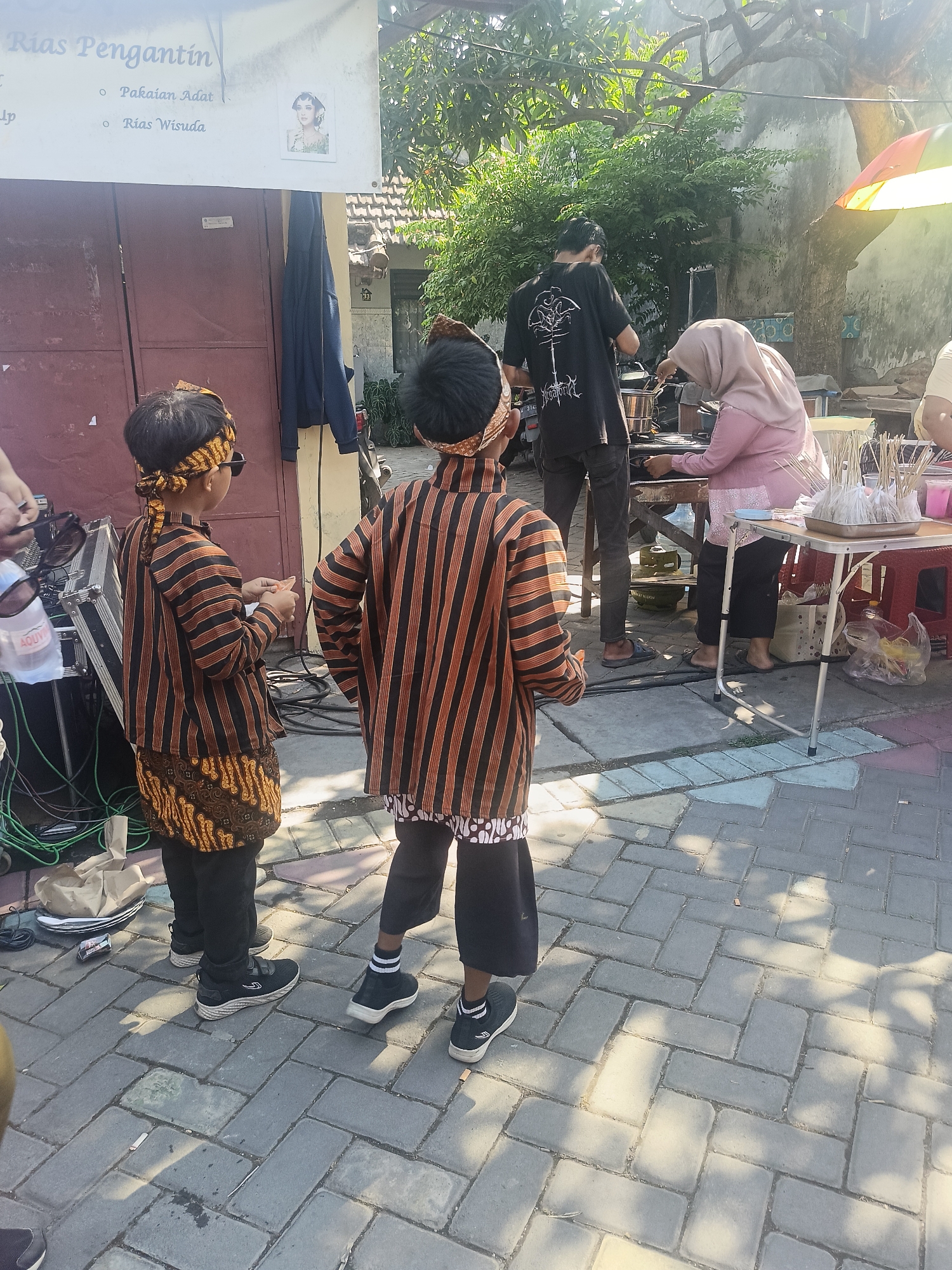 In Gedangan, Indonesia, two young boys dressed in traditional Javanese attire, specifically batik shirts with orange and black stripes and patterned waistcloths, stand facing away from the camera on a paved outdoor area. They both wear headbands and one is wearing sunglasses. Behind them, a man in a black t-shirt with a graphic design and dark pants is operating some equipment, possibly a sound system, as several cables are visible on the ground. To the right, a woman wearing a pink hijab and a pink top is attending to a table set up with various items, including skewers of food and drinks. The environment appears to be a street or an open-air market, with buildings and trees visible in the background, and a banner overhead advertising "Rias Pengantin" (Wedding Makeup) with images of traditionally dressed brides. The ground is paved with interlocking grey bricks, and sunlight casts shadows from trees and structures.