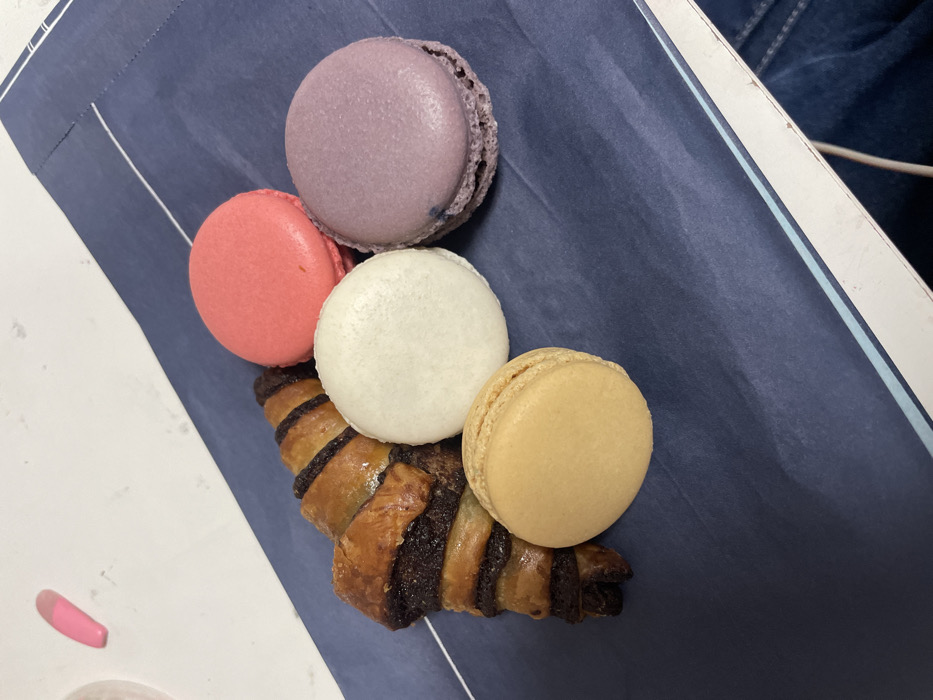 A collection of colorful macarons and a chocolate croissant are arranged on a blue surface. The macarons are pink, purple, white, and tan. The croissant is nestled beneath the white and tan macarons, showcasing its layered pastry and dark chocolate filling. The composition is set on a blue placemat, partially visible against a light background.