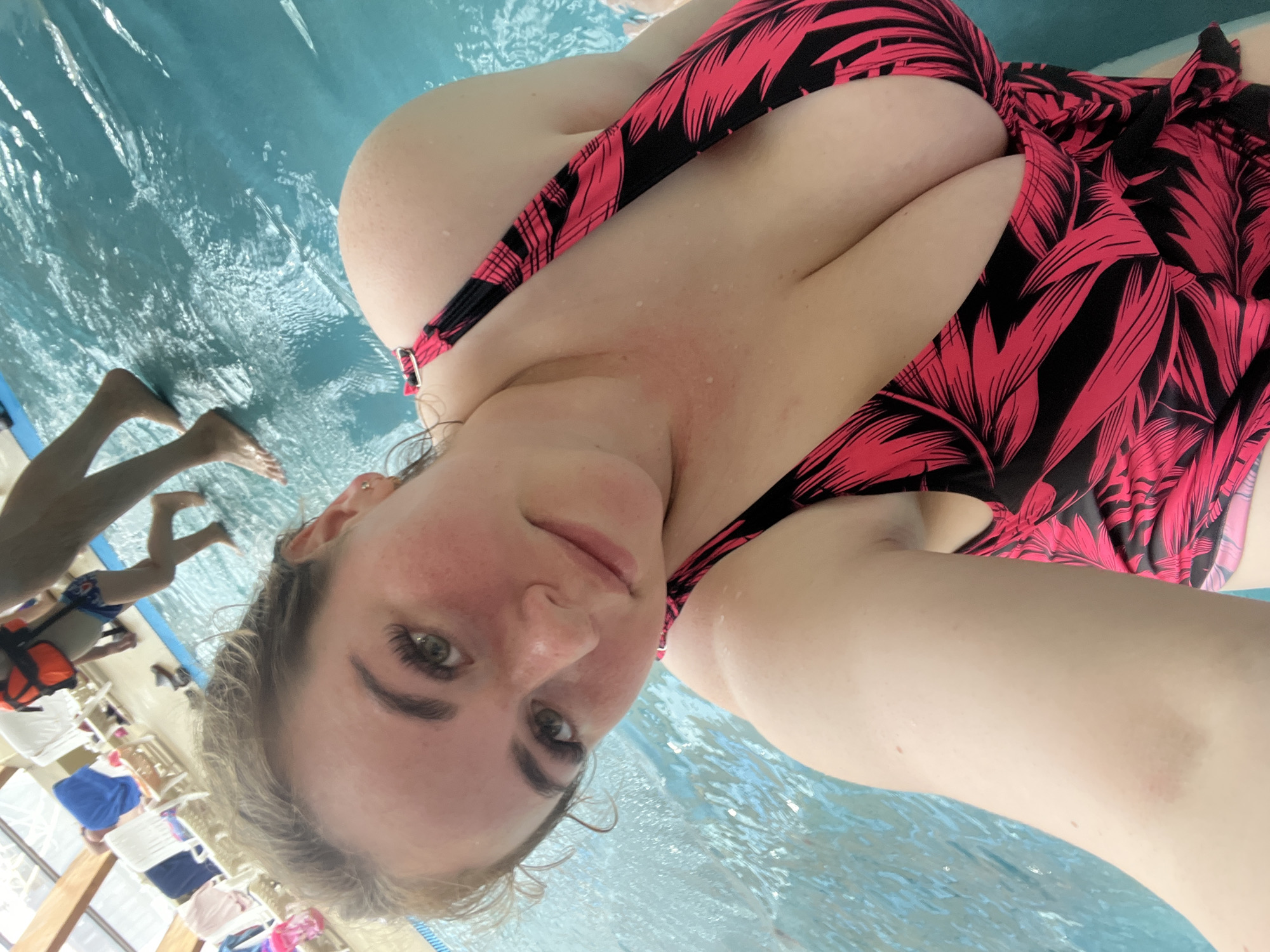 A woman with blonde hair, wearing a pink and black leaf-patterned bikini, takes a selfie from a swimming pool. She is looking towards the camera with a slight smile. Her eyes are green and her eyelashes are long and dark. The water in the pool is clear and blue, with some ripples visible. In the background, other people can be seen in the pool and around it. One person in the background is wearing blue and white swim trunks, and another is wearing a red life vest. There are lounge chairs and umbrellas visible in the background, suggesting an outdoor setting or a well-equipped indoor pool area. The lighting suggests it is daytime, possibly indoors given the presence of lounge chairs. The overall mood is relaxed and casual, typical of a day spent swimming.