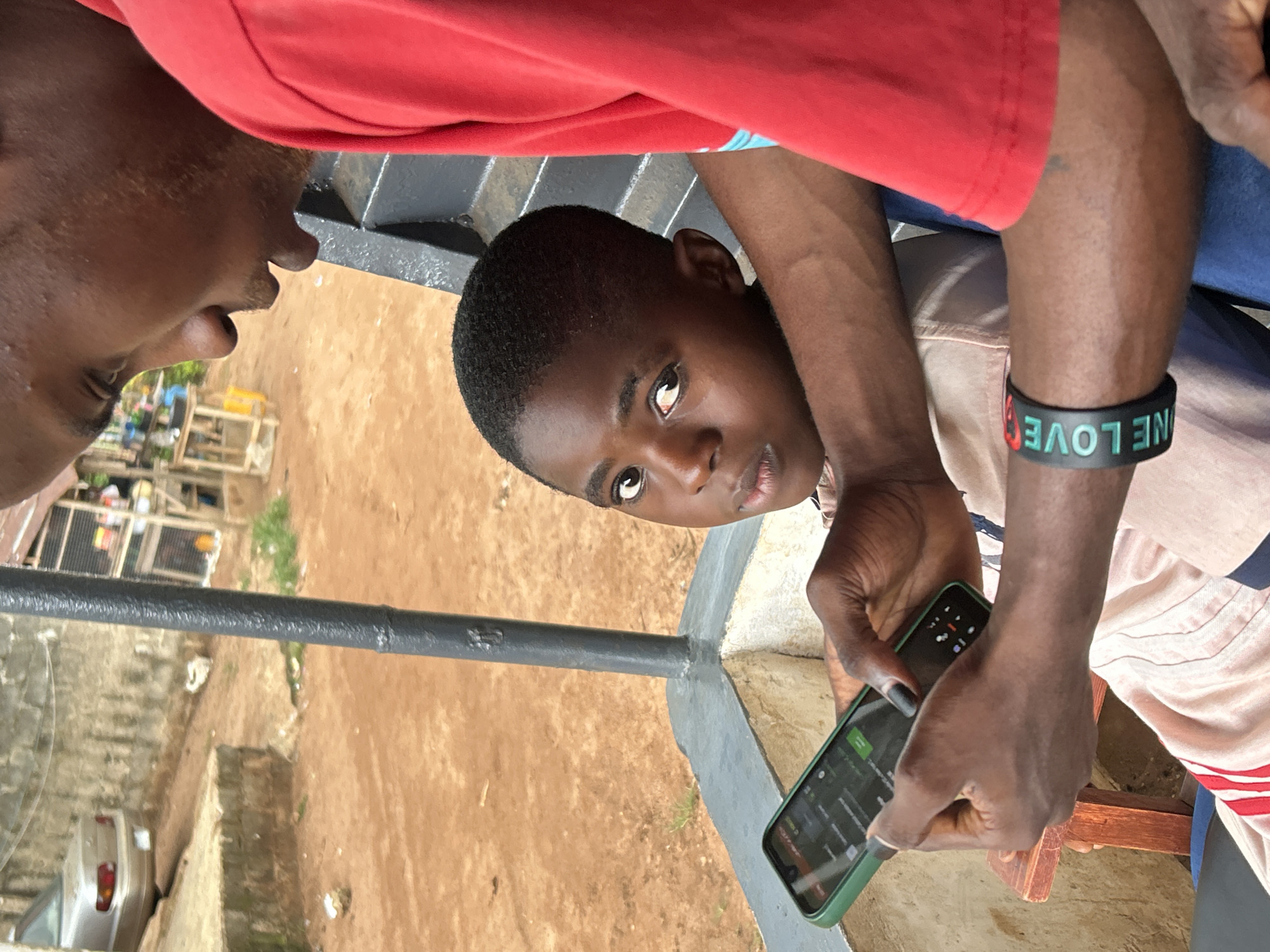 A medium, angled shot depicts a young boy holding a green smartphone with another person standing over his shoulder, presumably looking at the phone. The boy is dark-skinned, looking up with a slightly quizzical expression. A wristband that says "ONE LOVE" is visible on the adult's wrist. The environment shows a courtyard, with a gray pipe railing and reddish-brown dirt. In the background, there's a partial view of buildings, implying the location is an urban or semi-urban area. A silver car can be seen in the background.