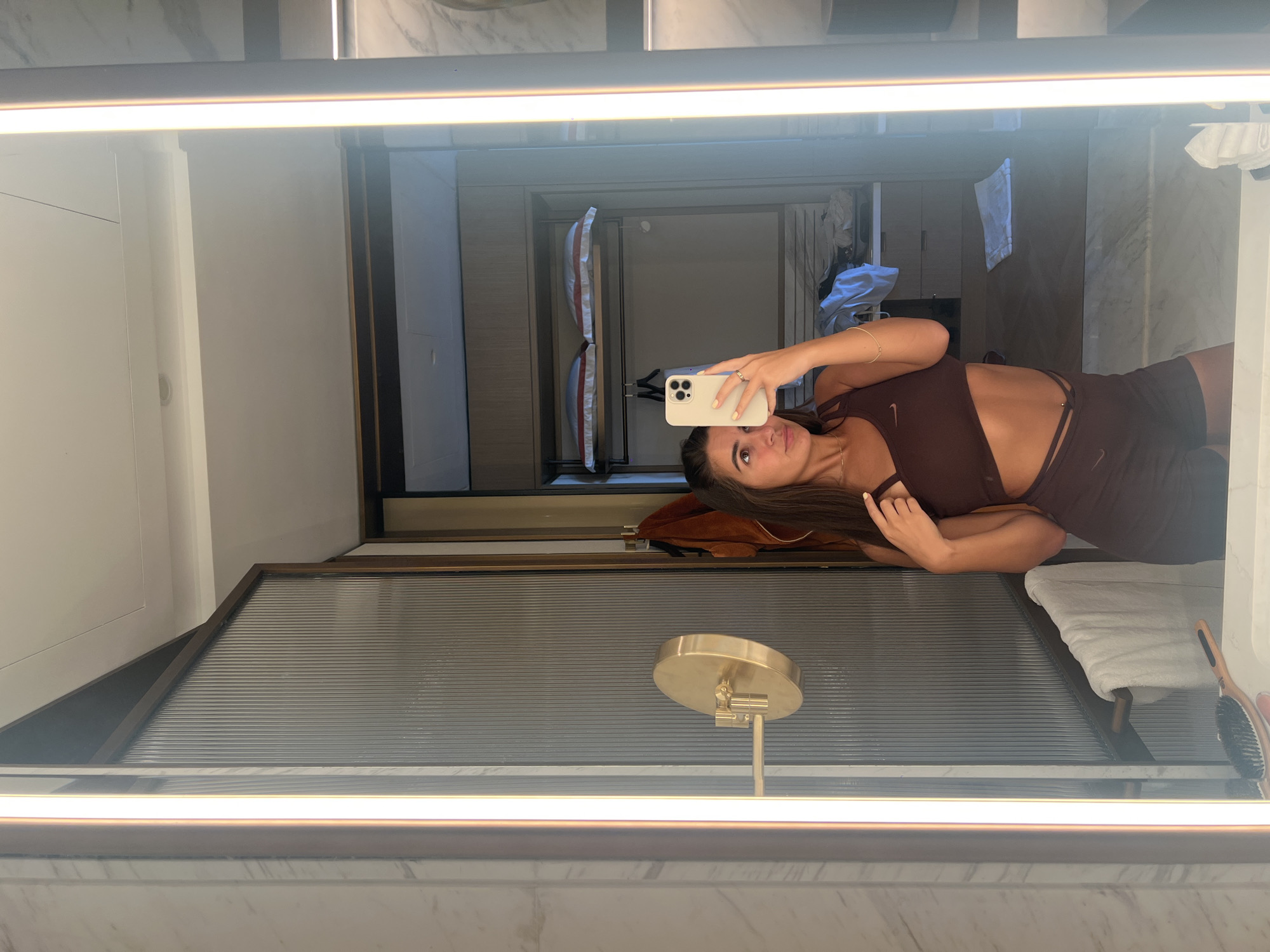 The image depicts a woman taking a mirror selfie inside a bathroom. The bathroom features marble surfaces and a vanity mirror with a light strip. In the reflection, a wardrobe with shelves is visible. A round, gold-colored vanity mirror is also present. The woman is wearing a dark maroon workout set. The location is likely a hotel or residential bathroom in Dubai, United Arab Emirates, given the modern interior design and luxurious finishes typical of Dubai properties.