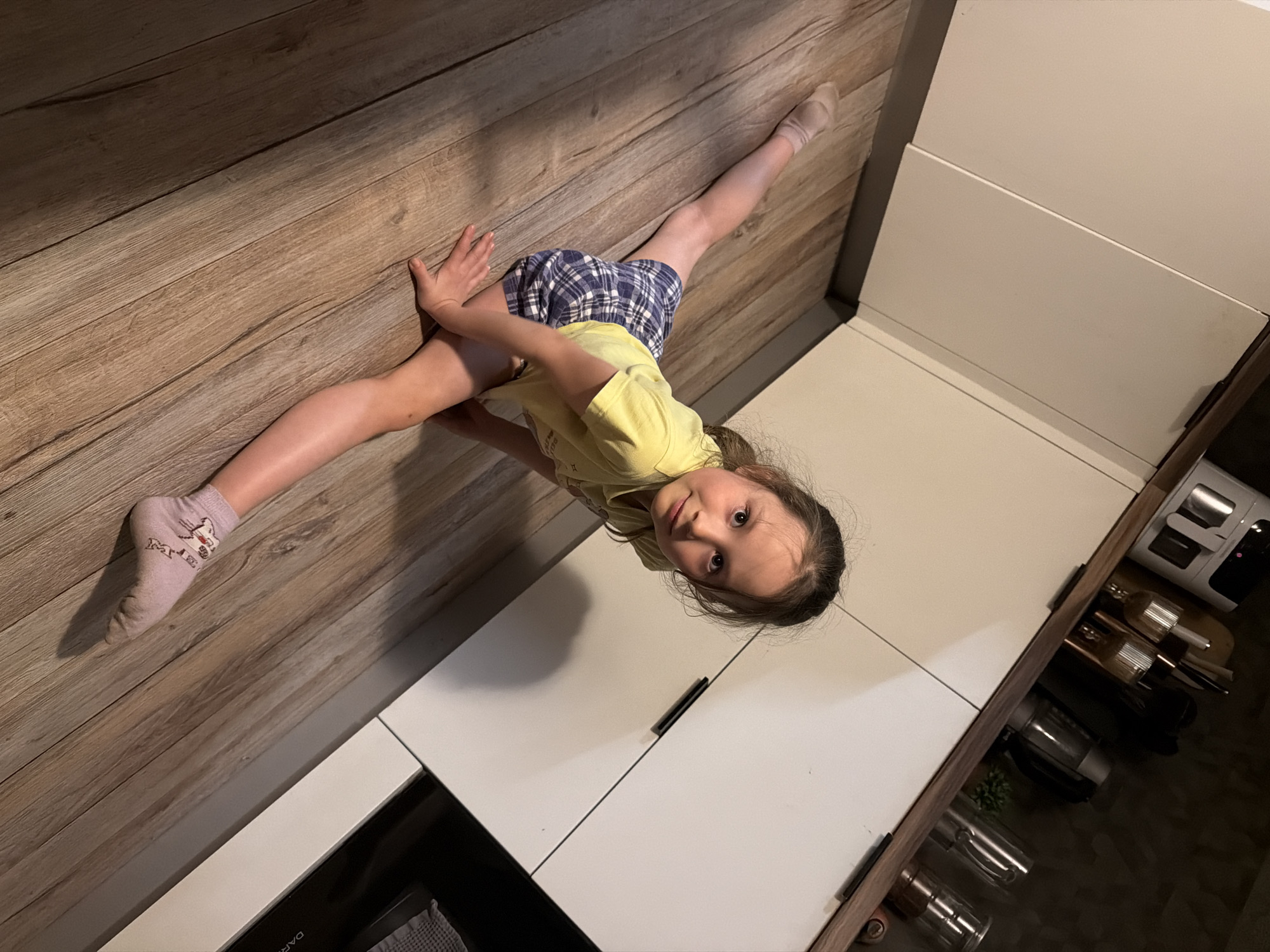 A young girl in a yellow t-shirt and blue and white plaid shorts is doing a split on a wooden floor next to white kitchen cabinets. She is wearing a sock on each foot. The location is likely inside a home, possibly in Perm, Russia, but there is no evidence to confirm the location. A drawer is partially open under the white cabinets.