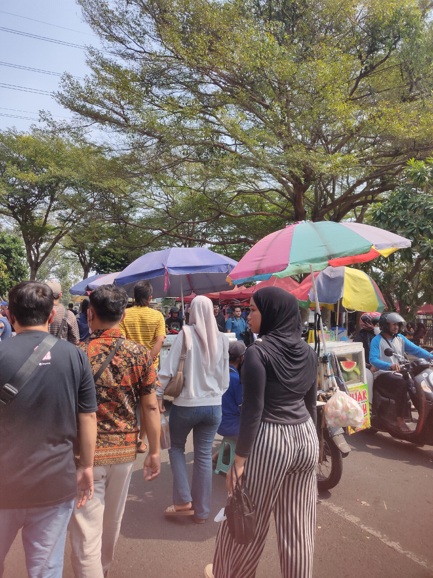 The image captures a lively outdoor market scene in Ngasem, Indonesia, under a clear, sunny sky. A crowd of people are navigating between stalls, many shaded by colorful umbrellas. In the foreground, the backs of several individuals are visible as they walk away from the camera. One person wears a black t-shirt, another a vibrant batik-patterned shirt with a red, orange, and black floral design, and a third, a person in a light-colored headscarf, is seen from the side. Further back, people in yellow and striped shirts are visible, as well as individuals browsing stalls. A food cart or stall displays a watermelon slice and other items, and a person on a scooter wearing a blue shirt and helmet is passing by. The environment is lush with trees providing a green canopy overhead.