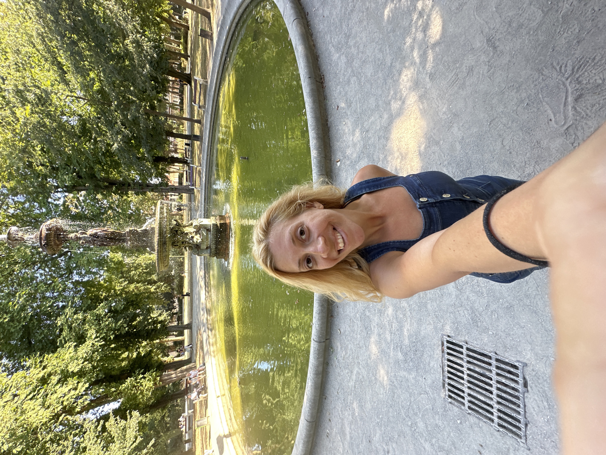 A woman with blonde hair is taking a selfie in front of a fountain in a park. She is wearing a denim overall dress. The fountain is circular with a tiered top and water flowing from it into a green pool. There are trees and benches around the fountain, and people can be seen in the background. The ground is paved with gravel and there is a grate on the left side of the image. The location is the Jardin du Fortifications in Calais, France.