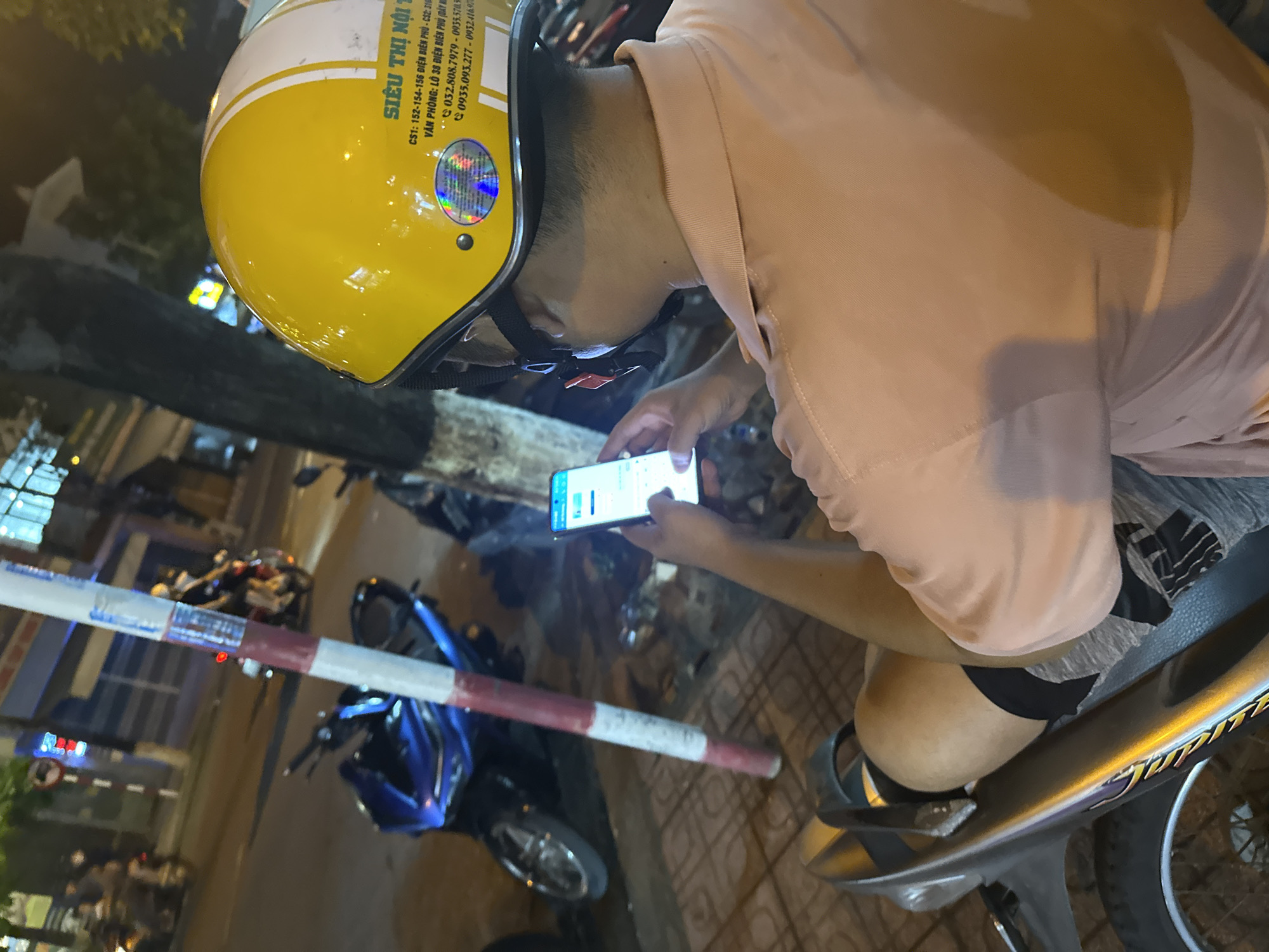A person wearing a yellow helmet and a light pink shirt is sitting on a moped. They are looking at their smartphone. A red and white barrier is visible in the foreground, and a blue motorcycle is parked nearby. In the upper left corner of the helmet, a holographic sticker with text that includes "SIÊU THỊ NỘI T" and phone numbers is partially visible.