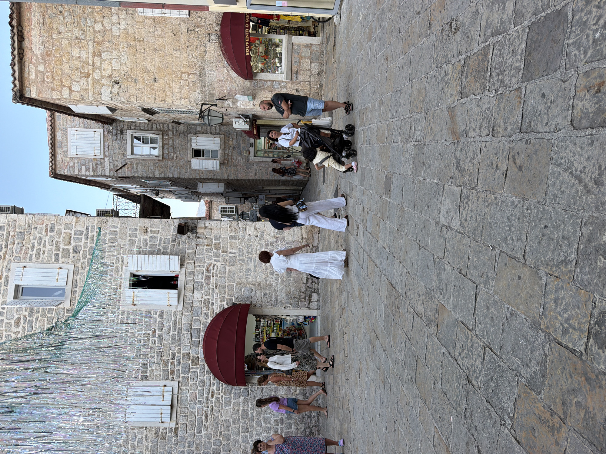 The image depicts a street scene in the Old Town of Budva, Montenegro. The architecture consists of stone buildings with traditional wooden shutters. In the foreground, a paved stone walkway is visible. Several people are present, walking and standing. A woman in a floral dress is on the left. Further into the scene, a group of individuals is seen walking past a building with a red awning. On the right side of the image, a man and a woman are standing near a stroller, with a young child partially visible. Another building on the right houses a souvenir shop. The overall atmosphere suggests a tourist area during daytime.