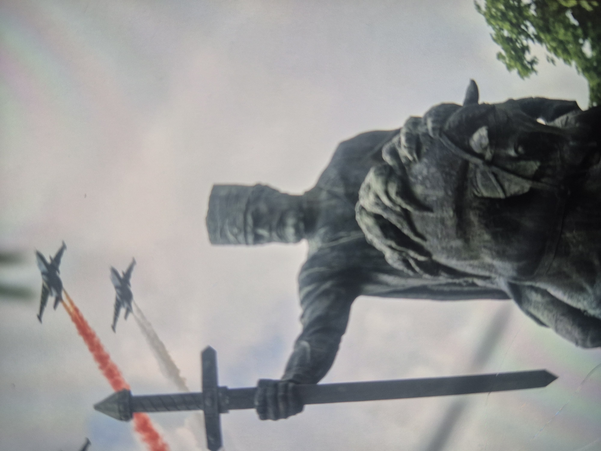 The image shows a statue of King Vakhtang Gorgasali on horseback, holding a sword, with two fighter jets flying above. The jets are trailing smoke in white and red. The statue is located in Tbilisi, Georgia. A tree is visible behind the statue. The sky is overcast.
