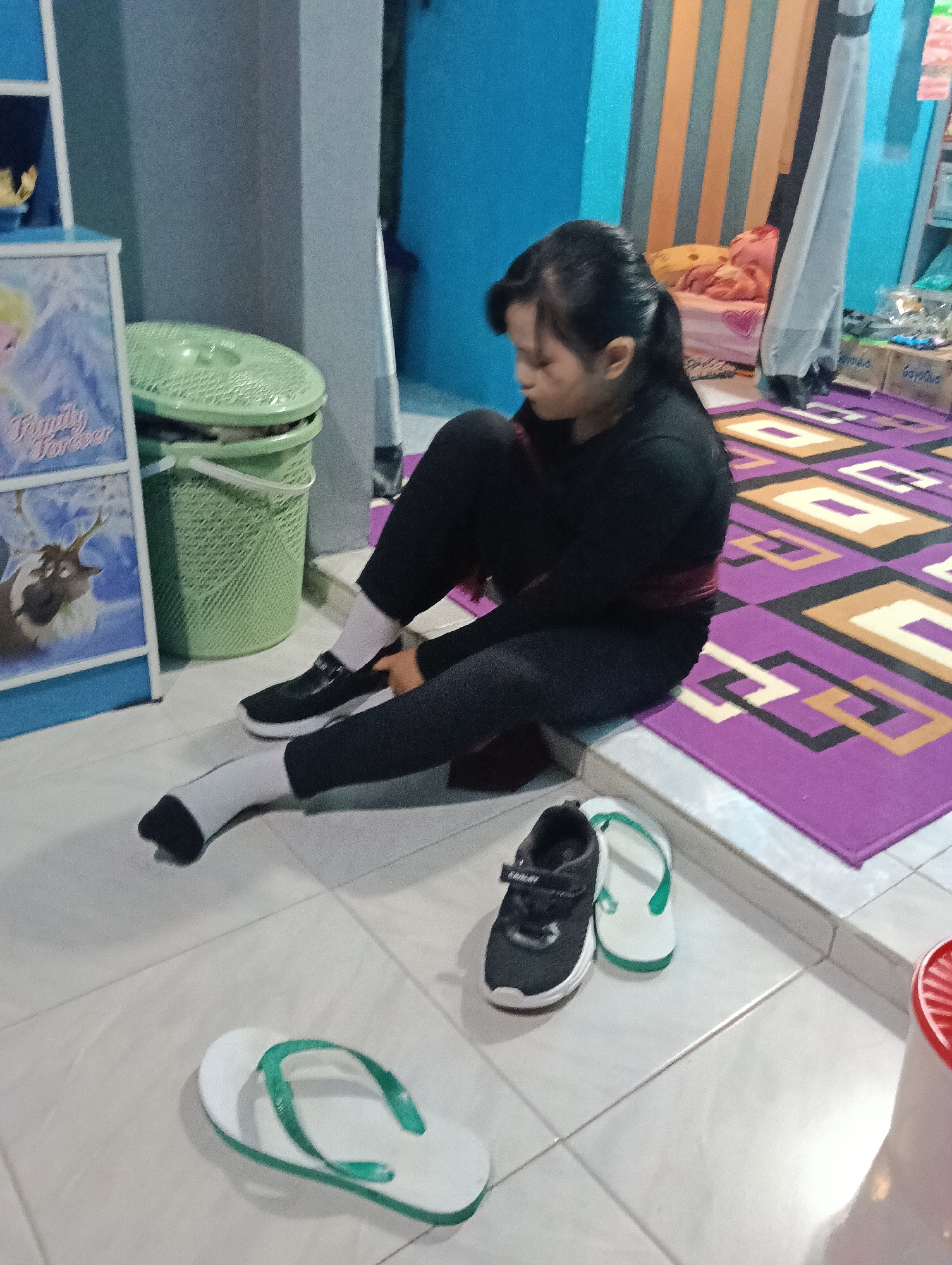 A young girl with her hair tied back in a ponytail is sitting on the floor, putting on black shoes with white soles and white socks. She is wearing black leggings and a black long-sleeved shirt with a red band around her waist. In front of her are a pair of white and green flip-flops and a pair of black sneakers with white soles and green accents. The floor is tiled with a light beige color. Behind her, there is a purple rug with a geometric pattern of purple, yellow, and white squares. A green laundry basket with a lid is visible to her left, next to a framed picture featuring characters from the movie "Frozen." The background shows blue and orange striped curtains and some shelves with items on them. The scene appears to be indoors, likely in a bedroom or play area. The lighting suggests it could be daytime. The overall mood is casual and domestic. The visible text on the framed picture reads "Family" and "Forever."