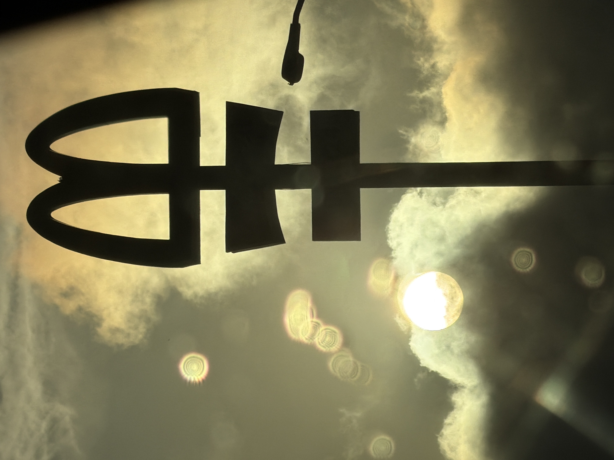 The image is a low-angle, upward-facing shot of a large, dark silhouette against a cloudy, bright sky. The perspective suggests the camera is looking up, possibly from below a sign or structure.
The central subject is a prominent, dark silhouette resembling a stylized emblem or logo. It appears to form the letters "BH" or a similar symbol. The left part is a rounded, open shape similar to the loop of a "B," while the right part is a more angular, block-like form, possibly an "H," connected to a long, horizontal bar extending to the right edge of the frame. This emblem is strongly backlit, highlighting its dark form against the bright background.
Above the emblem, a dark silhouette of what appears to be a street light or an overhead lamp is visible, hanging downwards.
The sky is filled with clouds, exhibiting warm, golden-yellow and lighter white tones, suggesting either sunrise or sunset. The overall lighting is very bright, creating a strong backlighting effect that renders the foreground objects as dark silhouettes. A distinct, bright circular light source, likely the sun, is visible in the lower right portion of the image, partially obscured by clouds. Several circular lens flares, some with concentric ring patterns, are scattered across the image, particularly in the lower half, indicating a direct shot into a strong light source.
No specific activity or event is taking place, and no people are visible. The image captures an atmospheric scene with a focus on light, shadow, and a prominent architectural or branded element. The stylized "BH" is the only discernible graphical information, which could be interpreted as text or a logo.