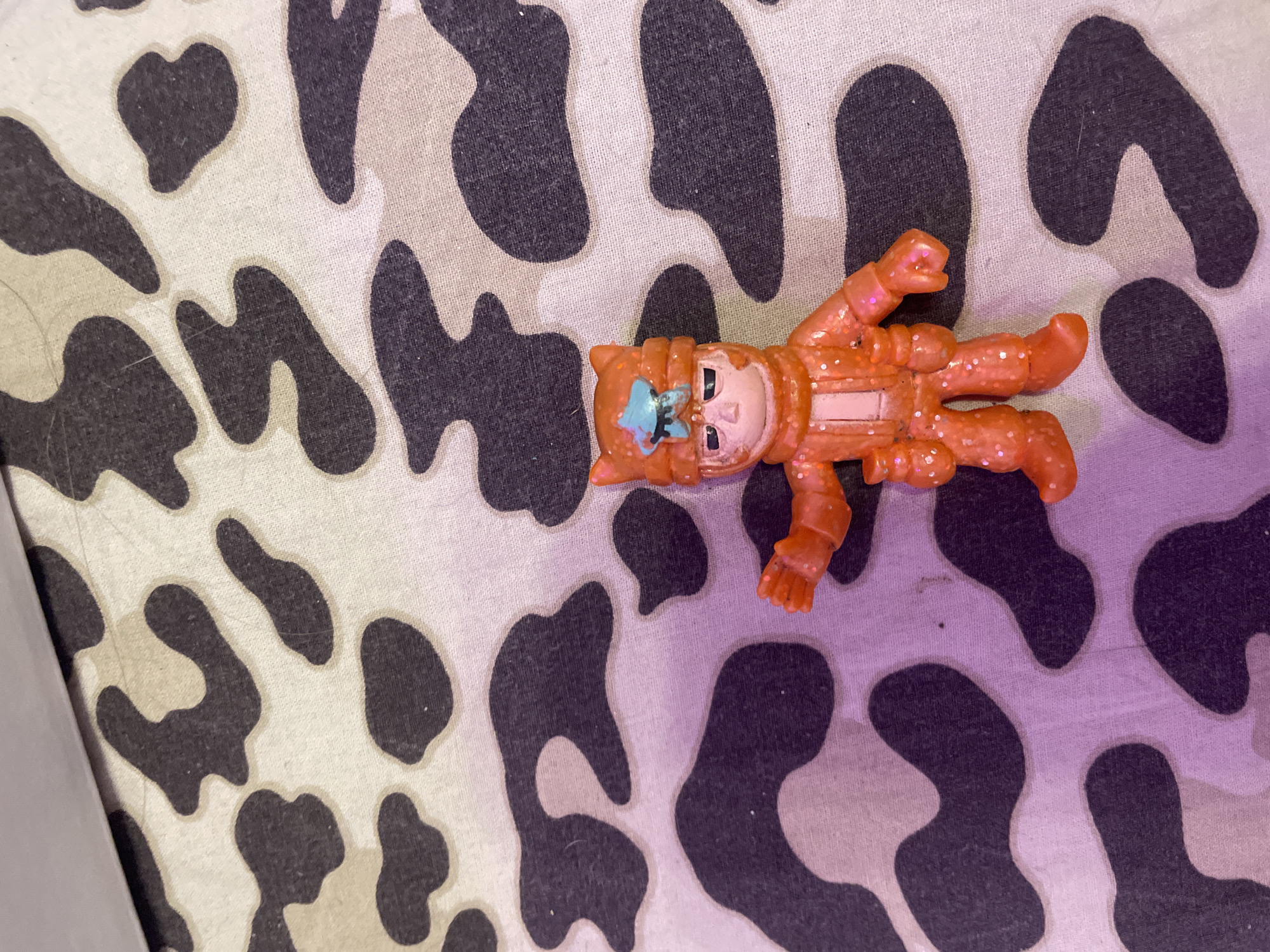 A small, orange, glitter-covered toy figurine of a character wearing a helmet with cat-like ears stands on a camouflage-patterned surface. The figurine has a blue star emblem on its helmet and appears to be a male character with a stern expression. The camouflage pattern consists of irregular black and light beige shapes. The lighting in the image has a purple hue, particularly visible on the lower left side. There is no information about the location in Vaganovo, Russia, provided in the image.