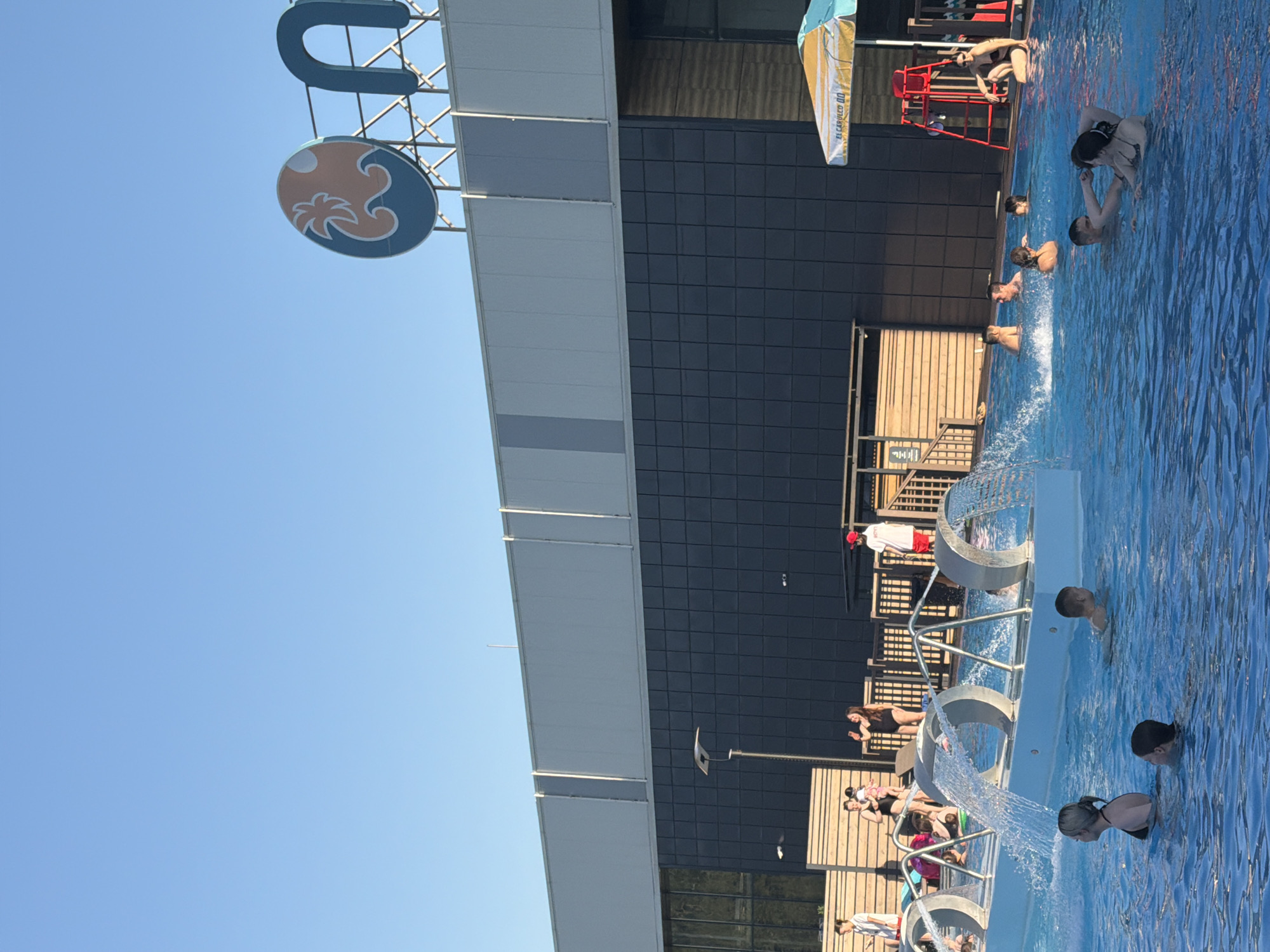 The image depicts a water park in Kazan, Russia, under a clear blue sky. The foreground shows a section of a pool with several people swimming and interacting with water features such as fountains. The pool is surrounded by wooden decking. A lifeguard stand with a lifeguard is visible. In the background is a building with a dark, tiled exterior and a white trim. A sign with a blue "U" and a circular emblem with a palm tree design is mounted on the building. There is a flag on a flagpole near the building.