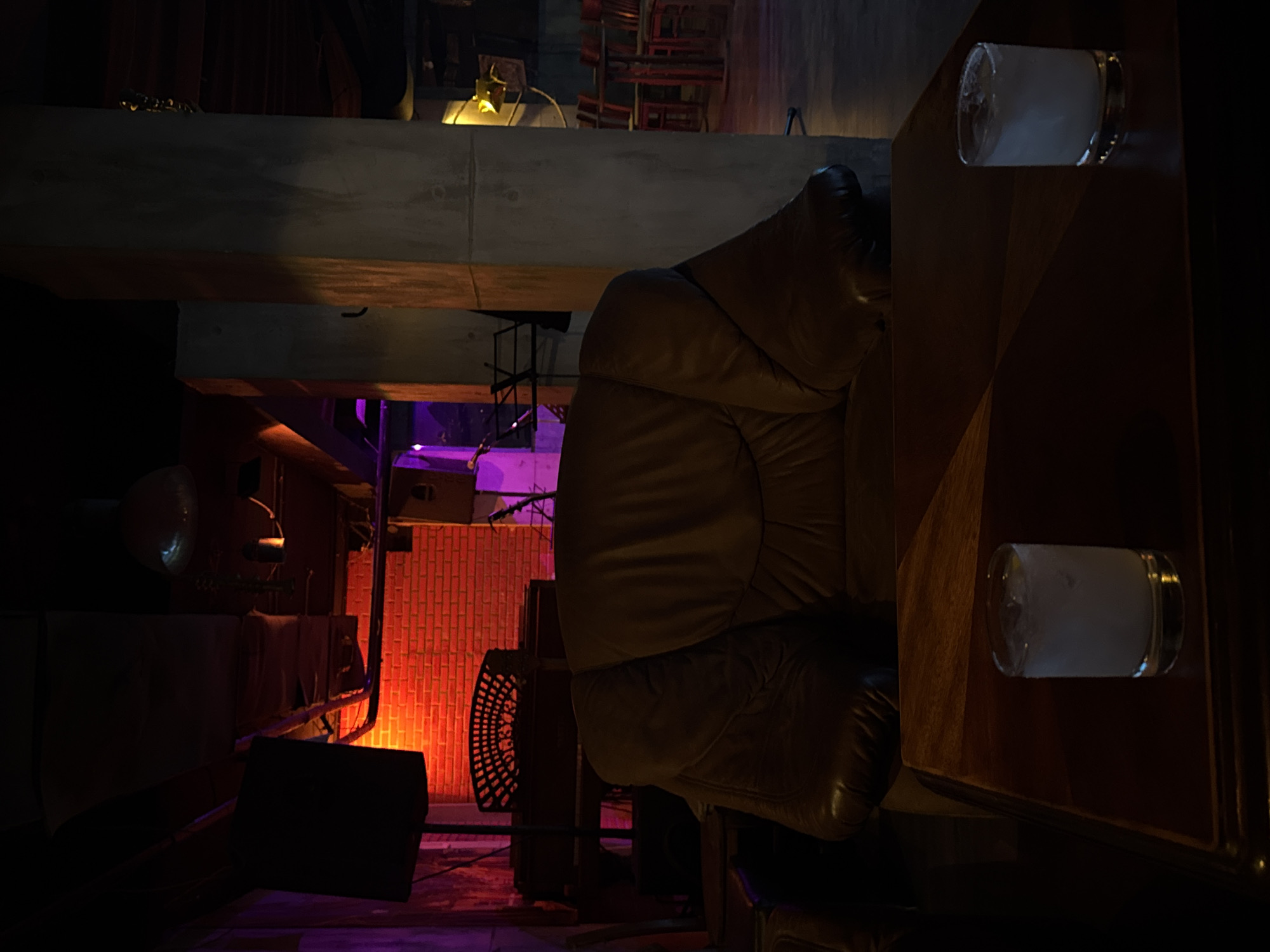 The image depicts the interior of a dimly lit bar, potentially in Sofia, Bulgaria, although there is no explicit evidence in the image confirming this location.
The primary subject is a large, brown, leather armchair, positioned centrally in the frame. To the right of the armchair is a wooden bar counter, with two glasses filled with a cloudy liquid resting on its surface.
In the background, a brick wall is illuminated with a reddish-orange light, possibly from an unseen source. A black speaker box sits on the floor, partially obscured by shadows. Above the armchair, a concrete support beam is visible. Red chairs are out of focus in the distance.
The environment suggests a relaxed, dimly lit atmosphere, typical of a bar or lounge. The focus is on the comfortable seating and the bar area, implying a social setting.