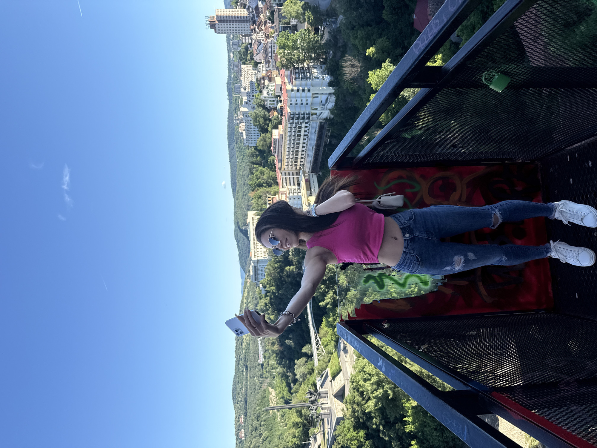 An outdoor, medium shot shows a young woman taking a selfie on a metal platform overlooking a city. She is wearing a pink crop top, distressed blue jeans, and white sneakers, with sunglasses on her head and a small purse over her shoulder. The woman is smiling and holding her phone up to take a picture. The background features a panoramic view of a city with buildings nestled among lush green hills, under a clear blue sky. A monument and a bridge are visible in the distance. The platform has a black metal railing with graffiti on the wall behind the woman.