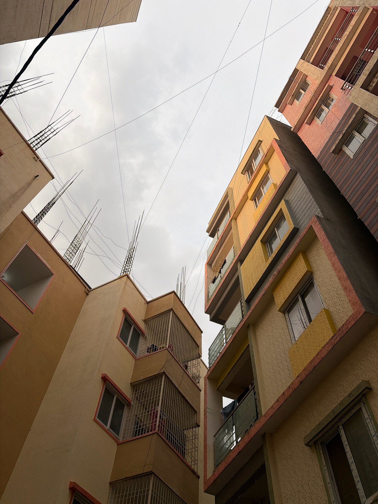 An upward-angle shot shows two multi-story apartment buildings in Bengaluru, India, under a cloudy sky. The building on the left is primarily cream-colored with some orange accents around windows and a square opening on the upper floors. Exposed rebar protrudes from the rooftop of this building, suggesting ongoing construction or recent work. Wires crisscross the sky between the buildings. The building on the right has a more colorful facade, with sections in shades of peach, yellow, and brown. It features balconies with glass railings and windows. The texture of the plaster on this building varies, with some areas exhibiting a raised patterned finish. The scene appears to be in a densely populated urban area of Bengaluru, likely a residential neighborhood. No people are clearly visible in the frame, but some objects suggest occupancy, such as laundry hanging on a balcony. The lighting is soft due to the overcast sky.