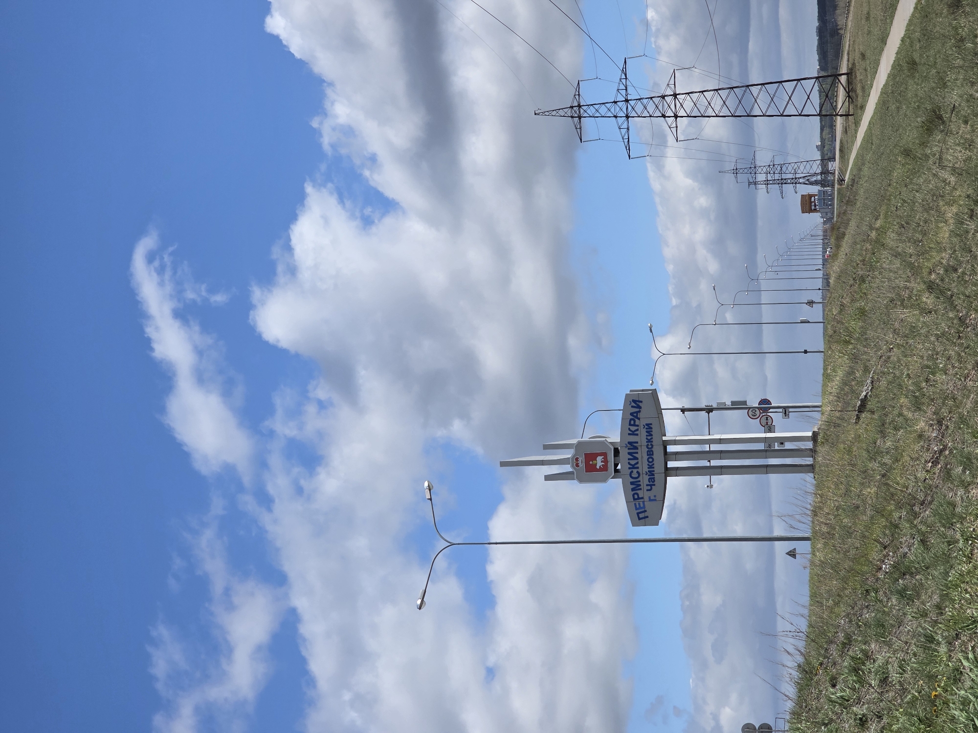 The image shows an outdoor scene in Noviy, Russia, featuring a blue sky with scattered white clouds. An electrical pylon with wires stretches across the top of the frame. A roadside sign reads "Пермский край" (Perm Krai) and "г. Чайковский" (Chaykovsky City). Streetlights line the road, and a grassy verge extends along the right side of the frame. The overall setting appears to be a rural or suburban area at the edge of the city.