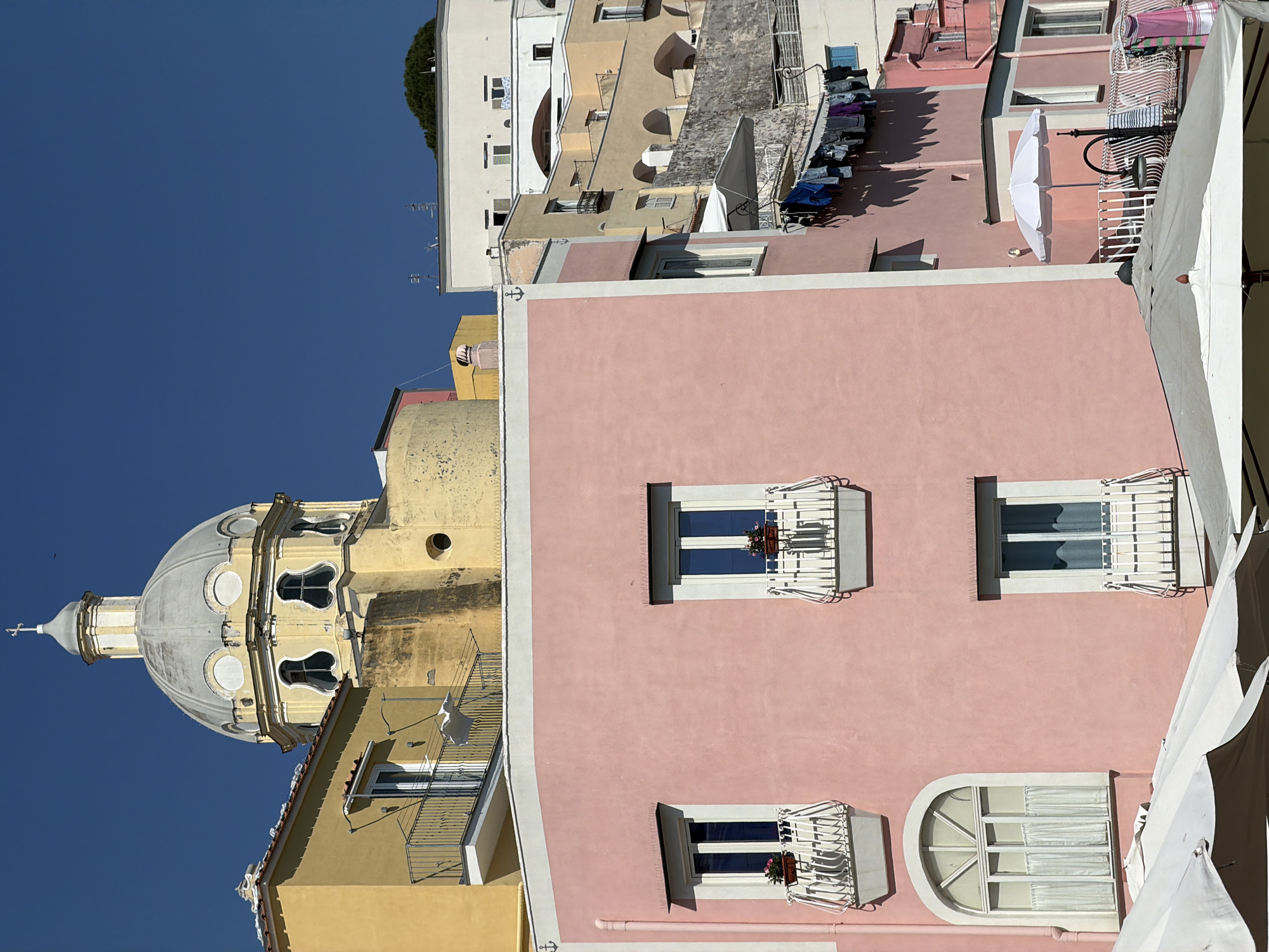 The image depicts a building in Procida, Italy. The structures are diverse, painted in colors such as pink, yellow, and white. The buildings are close together, some with balconies and windows adorned with flowers. A prominent feature is a large, dome-shaped church. The sky is clear and blue. The image also shows various architectural details like roofs, and decorative elements. Laundry is hanging out to dry on the roof of one of the buildings.