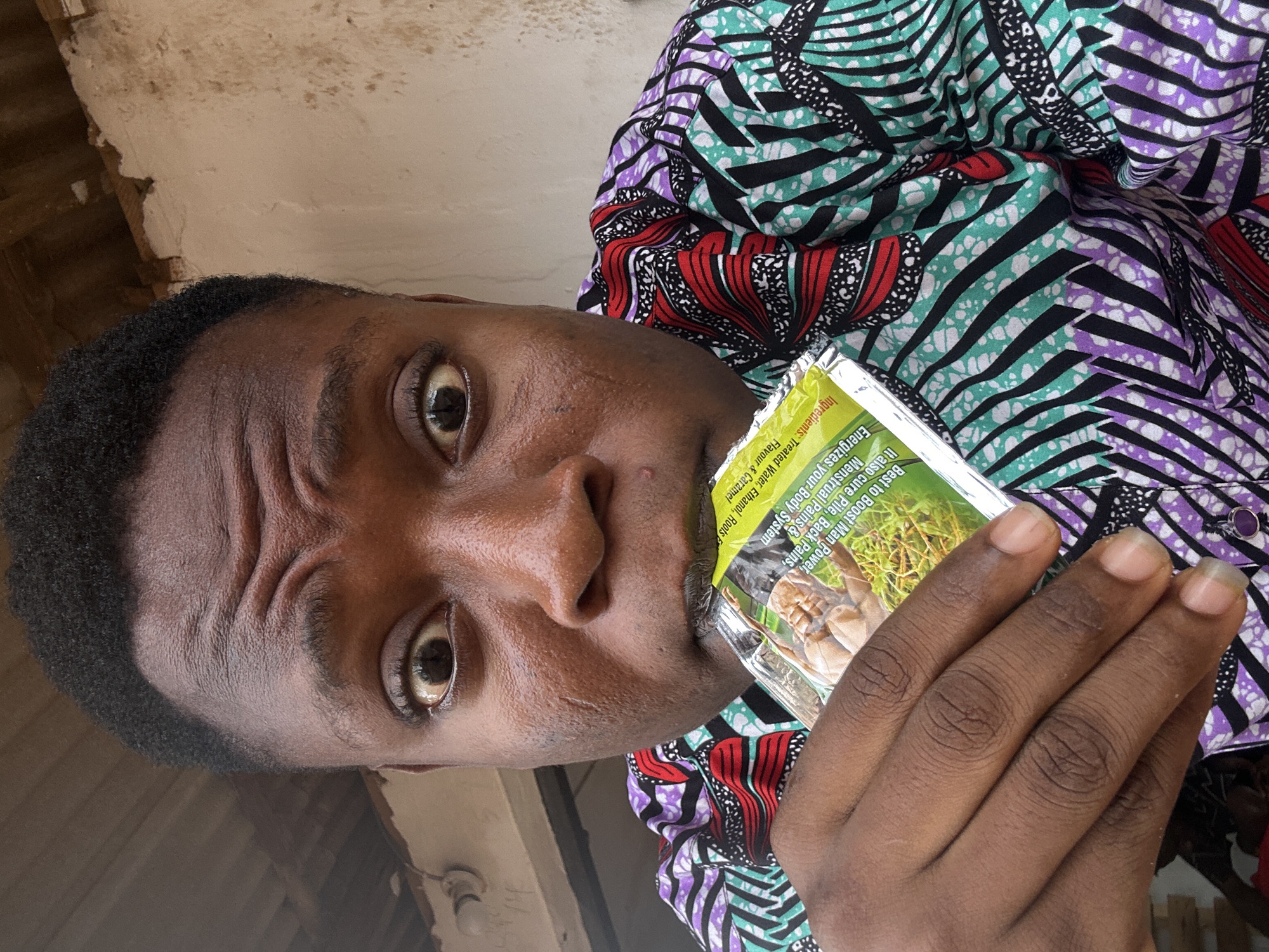 A selfie captures a person with dark skin and short, dark hair, holding an opened packet of what appears to be a health supplement in their mouth. The packet, mostly obscured by the person's face and hand, has text on it that reads "Best to Boost Man Power, Menstrual Pains & It also cure Pile, Back Pains, Energizes your Body System Ingredients: Treated Water, Ethanol, Roots & Flavour & Caramel." The person is wearing a colorful shirt with repeating patterns of green, black, purple, and red. The background appears to be a dilapidated building with exposed ceiling beams. The location is Unknown, Unknown.