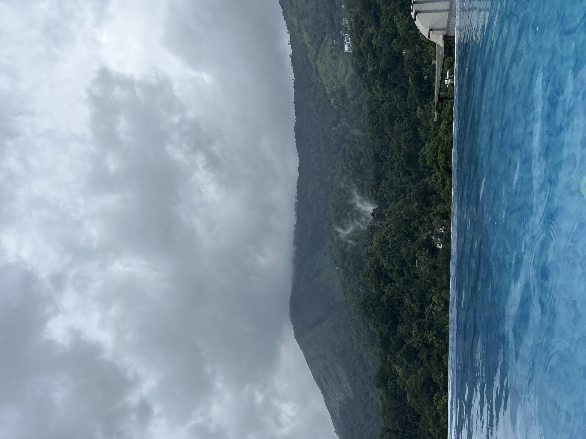 This image captures an expansive view of a mountainous, forested landscape in Anachal, India, beneath a heavily overcast sky, with an infinity pool in the foreground. The lower approximately 20% of the frame features a blue-tiled infinity pool, its surface displaying ripples that reflect the ambient light. The pool's edge creates a sharp, horizontal division across the image. Above the pool, the midground and background are dominated by dense, tropical forests covering rolling hills and imposing mountains.
Mist or low-lying clouds are visibly weaving through the trees on the mountain slopes, particularly concentrated on the left side of the midground. Scattered within the dense foliage are a few small structures; a distinct multi-story building with light-colored walls is visible on a distant, forested hill towards the right side of the frame. A dark, angular structure, possibly part of a viewing platform or resort infrastructure, is partially visible protruding from behind the pool's edge on the far right. A transmission tower structure is also partially visible among the trees on the right. The upper approximately 40% of the image is filled with a thick canopy of grey, diffused clouds, indicating an overcast day without direct sunlight. No people are present in the scene.