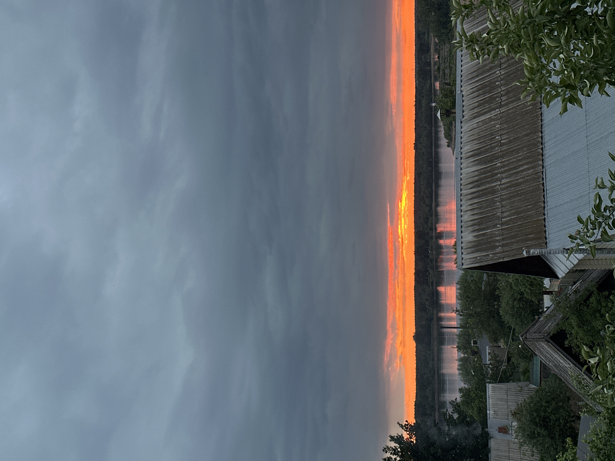 The image depicts a serene sunset over a river in Chaikovsky, Russia. The sky is filled with dramatic, dark gray clouds, juxtaposed against a vibrant band of fiery orange and yellow light near the horizon where the sun is setting. This colorful glow reflects on the calm surface of a wide river, creating shimmering streaks of light.
In the foreground, a rural settlement is visible. Several houses with corrugated metal roofs are clustered together. One house in the lower left has smoke emanating from its chimney. Lush green trees and foliage surround the buildings and line the riverbanks. The overall atmosphere is peaceful and tranquil, characteristic of a rural sunset.