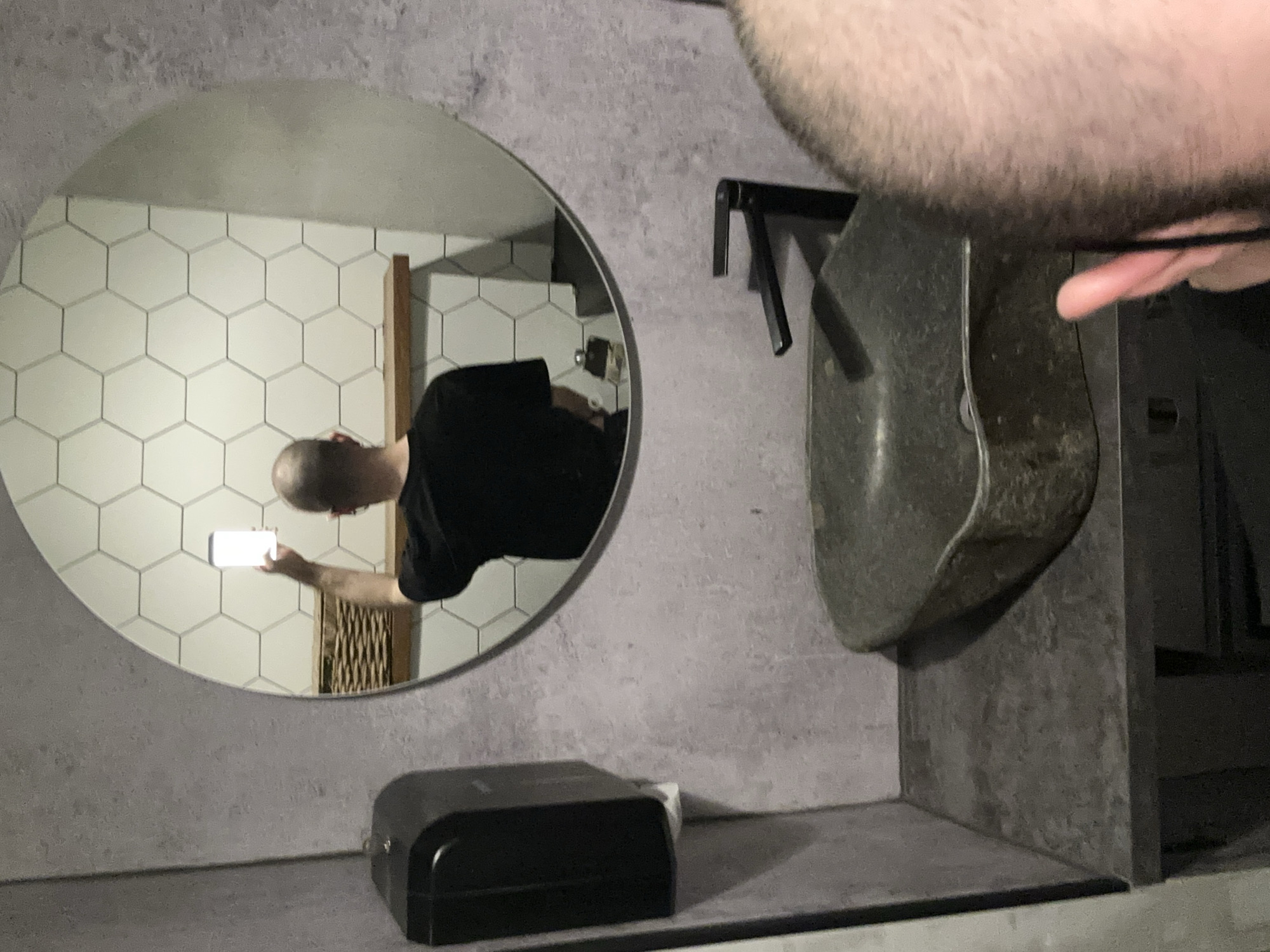 A man is taking a selfie in a bathroom. He is reflected in a round mirror on a wall covered in a gray material that resembles concrete. Behind the man in the mirror, the bathroom wall is covered in white hexagonal tiles. To the right of the mirror is a gray stone-like sink. There is a black soap dispenser on the counter below the mirror. There is no indication that this is Moscow, Russia.